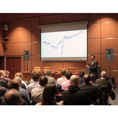 Ein Mann hält einen Vortrag vor Publikum in einem Konferenzraum. Ein Diagramm mit steigendem Trend ist auf der Leinwand zu sehen.