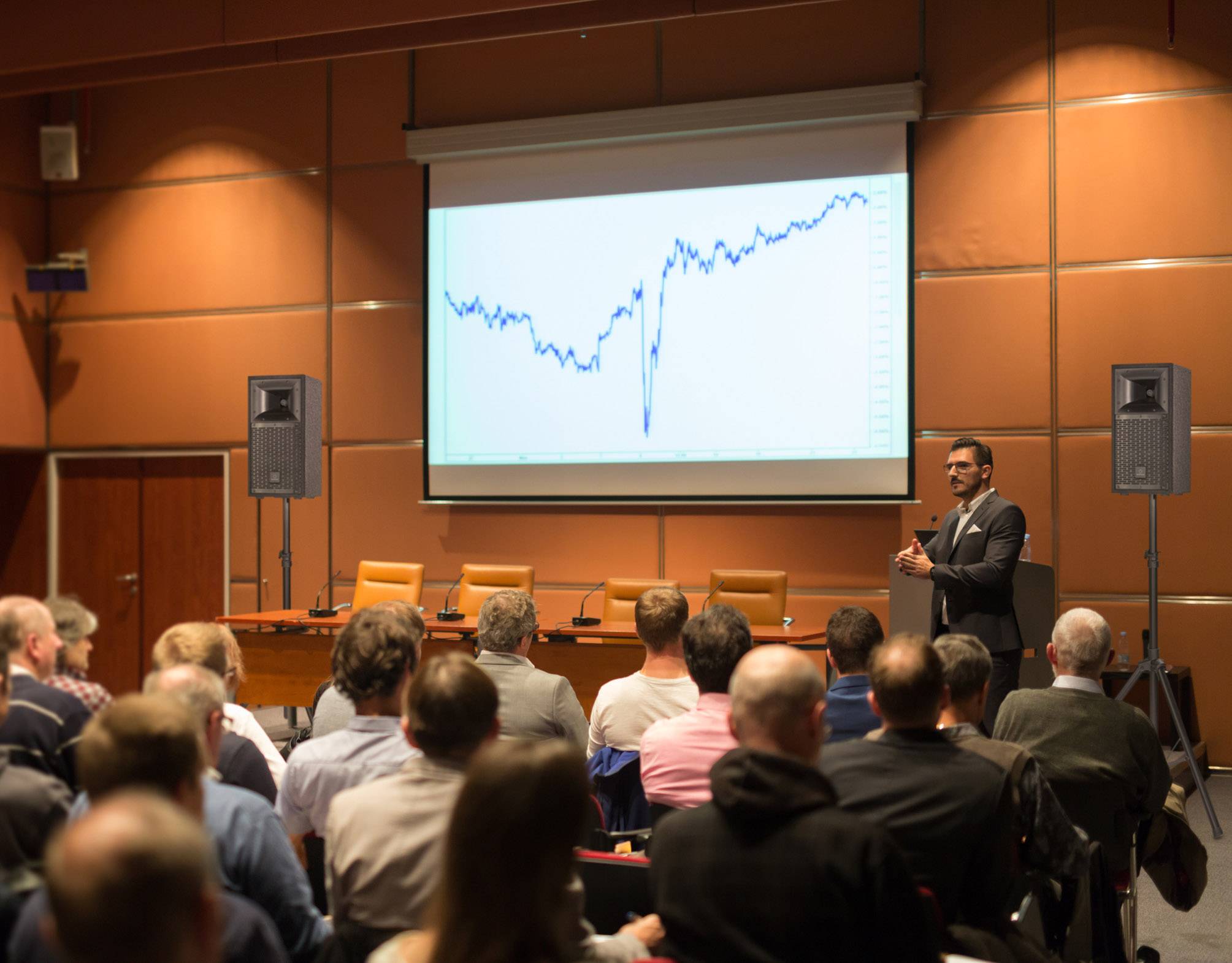 Person hält Vortrag vor Publikum in Konferenzraum. Im Hintergrund großes Diagramm mit Kursverlauf auf Leinwand. Thema: Aktienkursentwicklung.