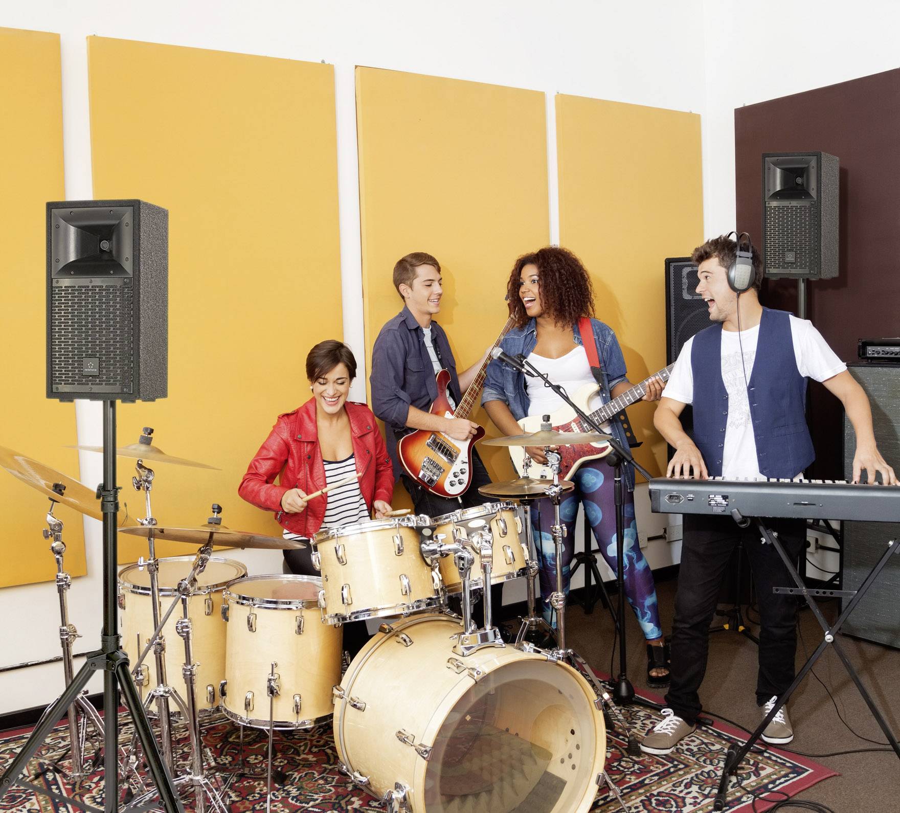 Vier Personen spielen in einem Musikproberaum. Eine Person spielt Schlagzeug, zwei spielen Gitarre und eine spielt Keyboard. Alle wirken fröhlich.