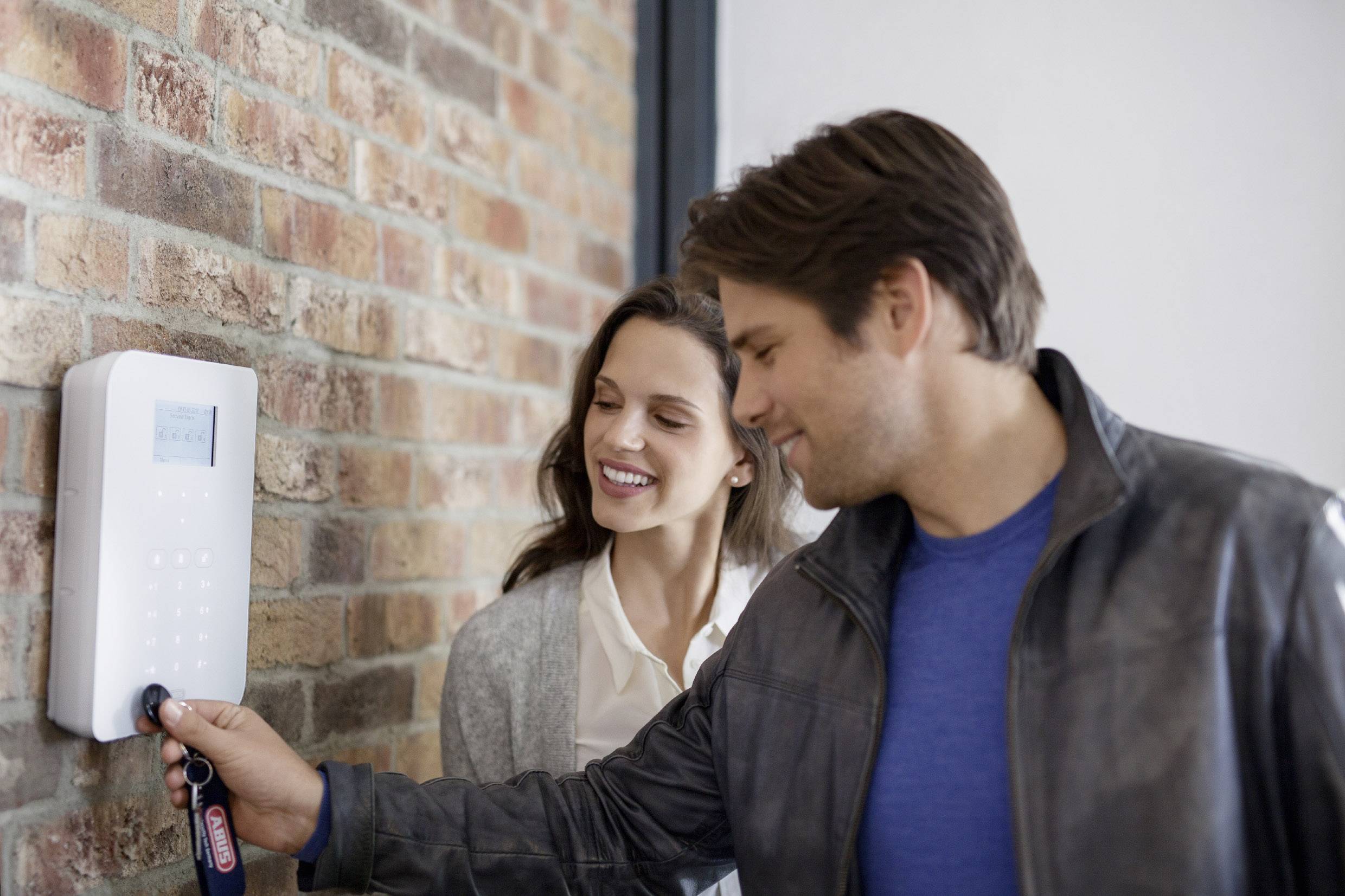 Ein Mann und eine Frau bedienen ein Sicherheitsalarmsystem an einer Backsteinwand. Der Mann hält einen Schlüsselanhänger an das System.