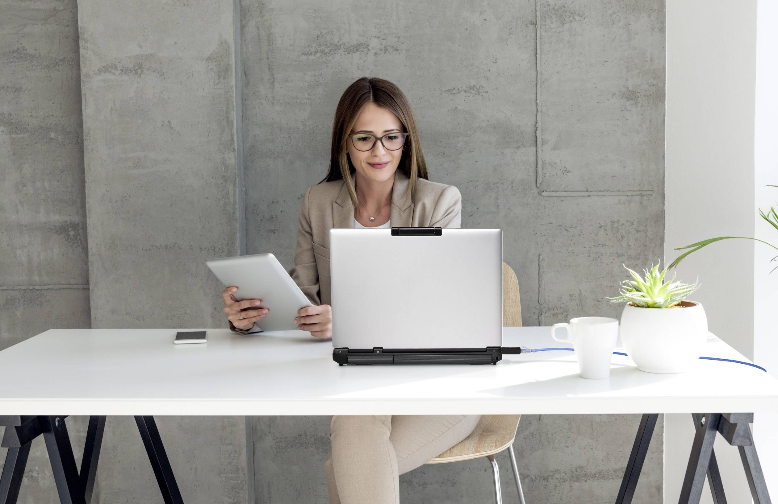Eine Frau arbeitet an einem Laptop am Schreibtisch. Sie hält ein Tablet und hat eine Kaffeetasse und eine Pflanze neben sich.