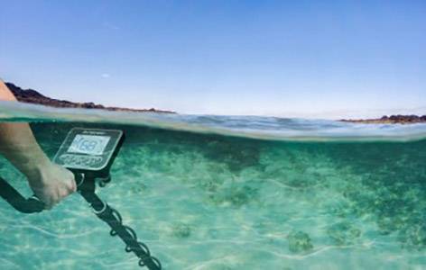Person verwendet einen Metalldetektor im flachen, klaren Meerwasser, umgeben von entfernten Hügeln am Horizont.