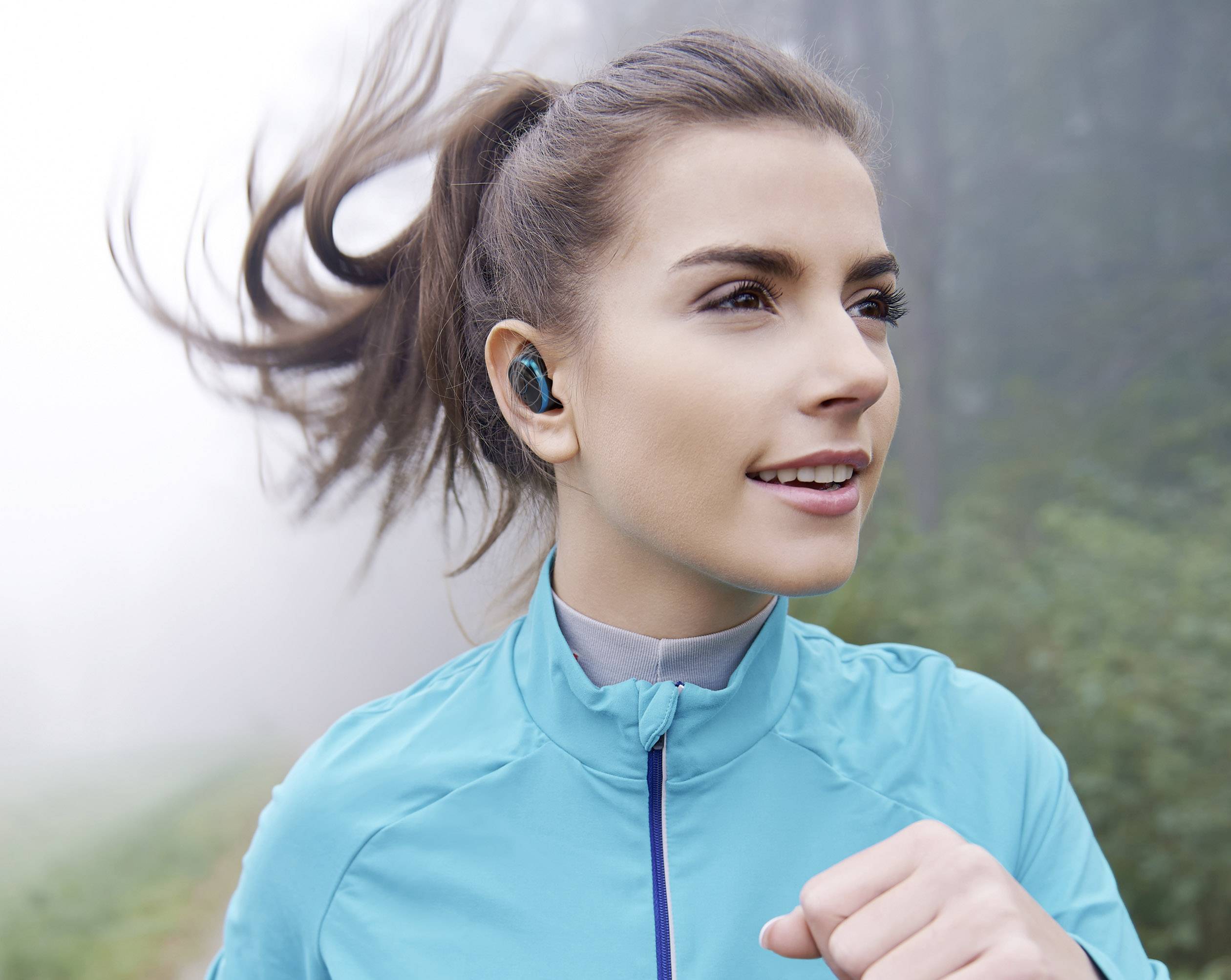Frau joggt in blauer Jacke an einem nebligen Tag. Sie trägt kabellose Ohrhörer und schaut konzentriert nach vorne.