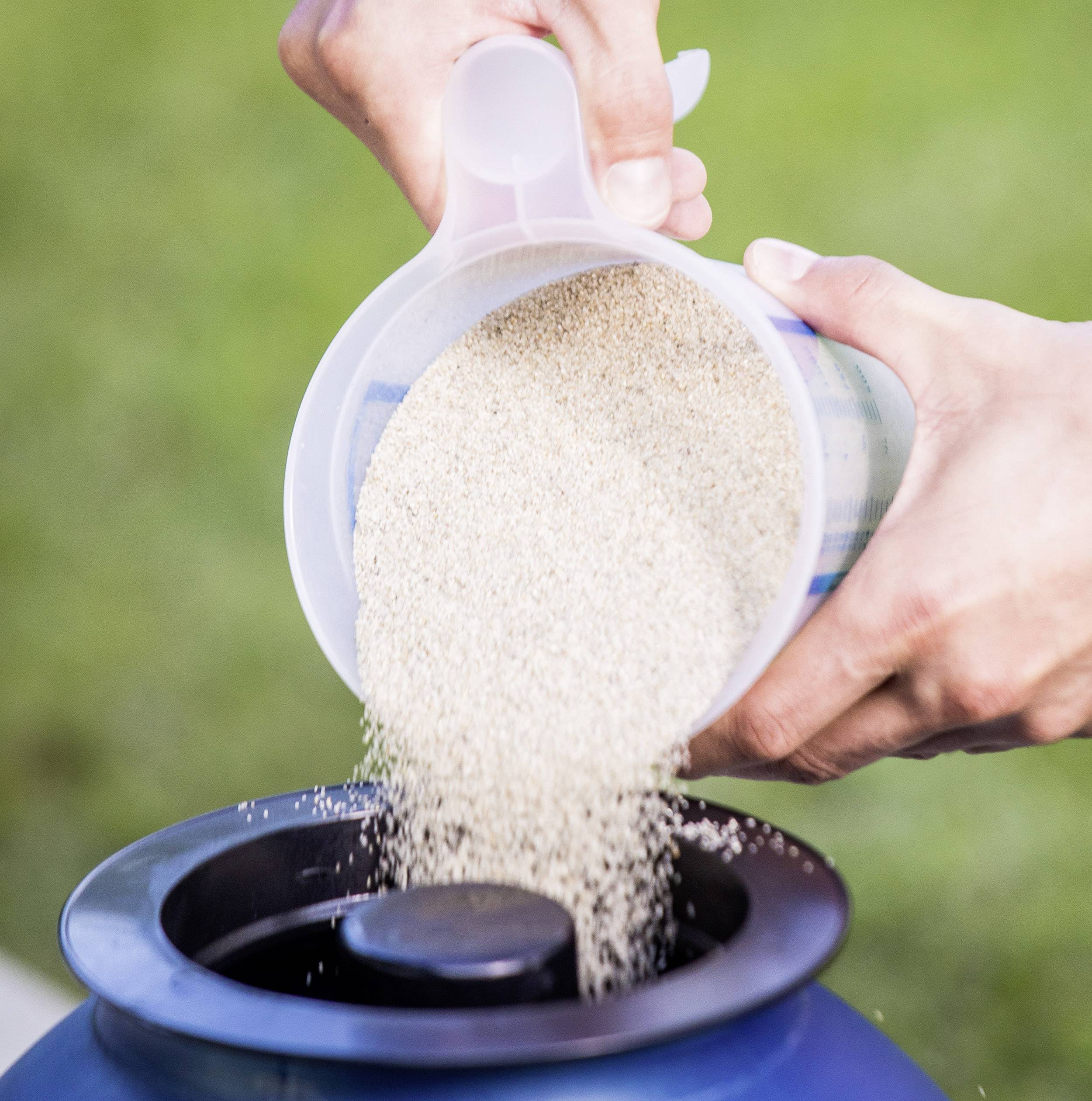 Person schüttet feines Granulat aus einem Messbecher in einen Behälter im Freien.