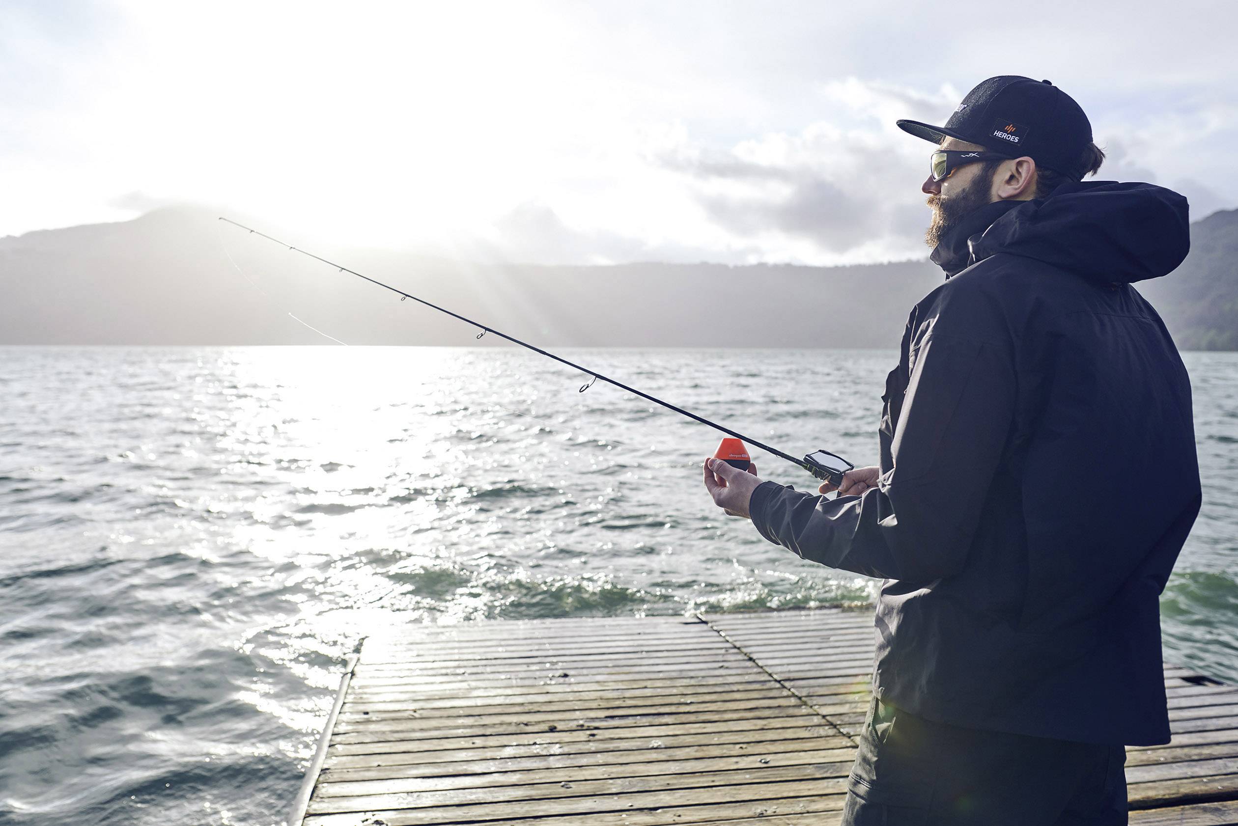 Eine Person angelt auf einem Steg am See bei Sonnenuntergang, trägt ein dunkles Outfit und eine Sonnenbrille.