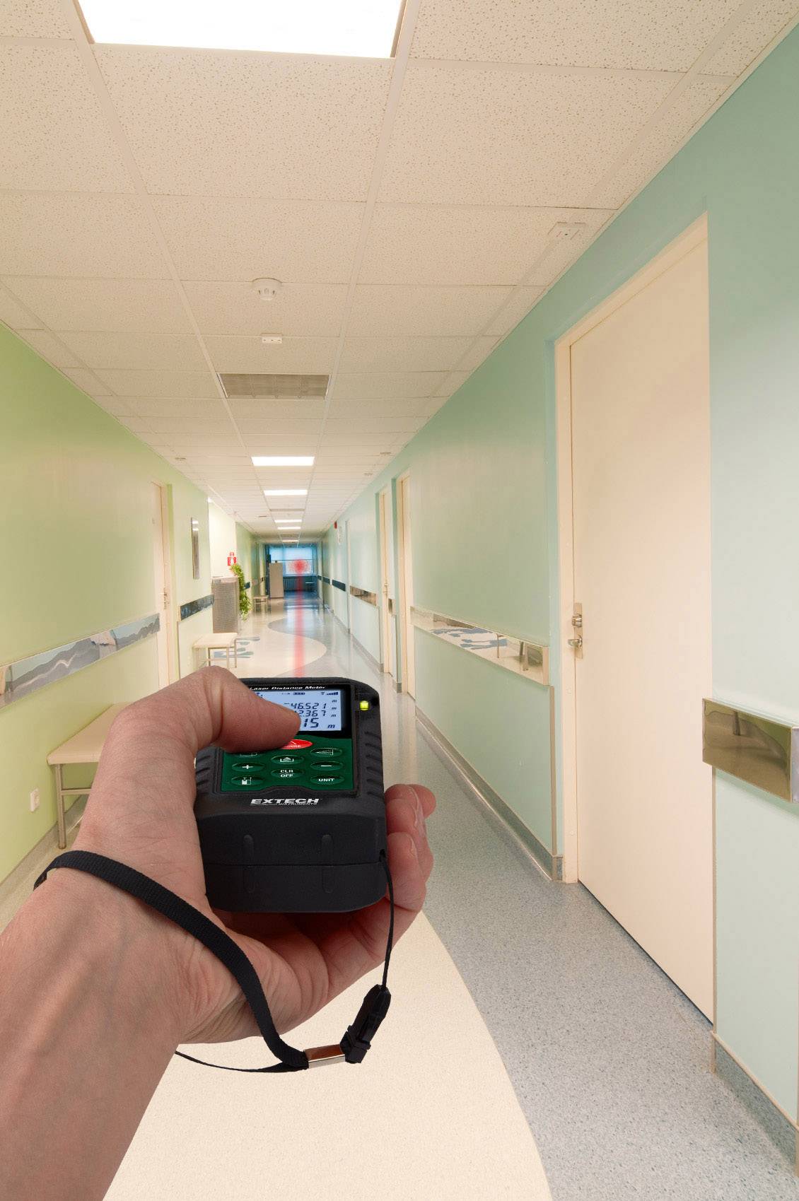 Eine Hand hält ein Laser-Entfernungsmessgerät in einem langen, hell erleuchteten Korridor mit grünen Wänden und geschlossenen Türen.