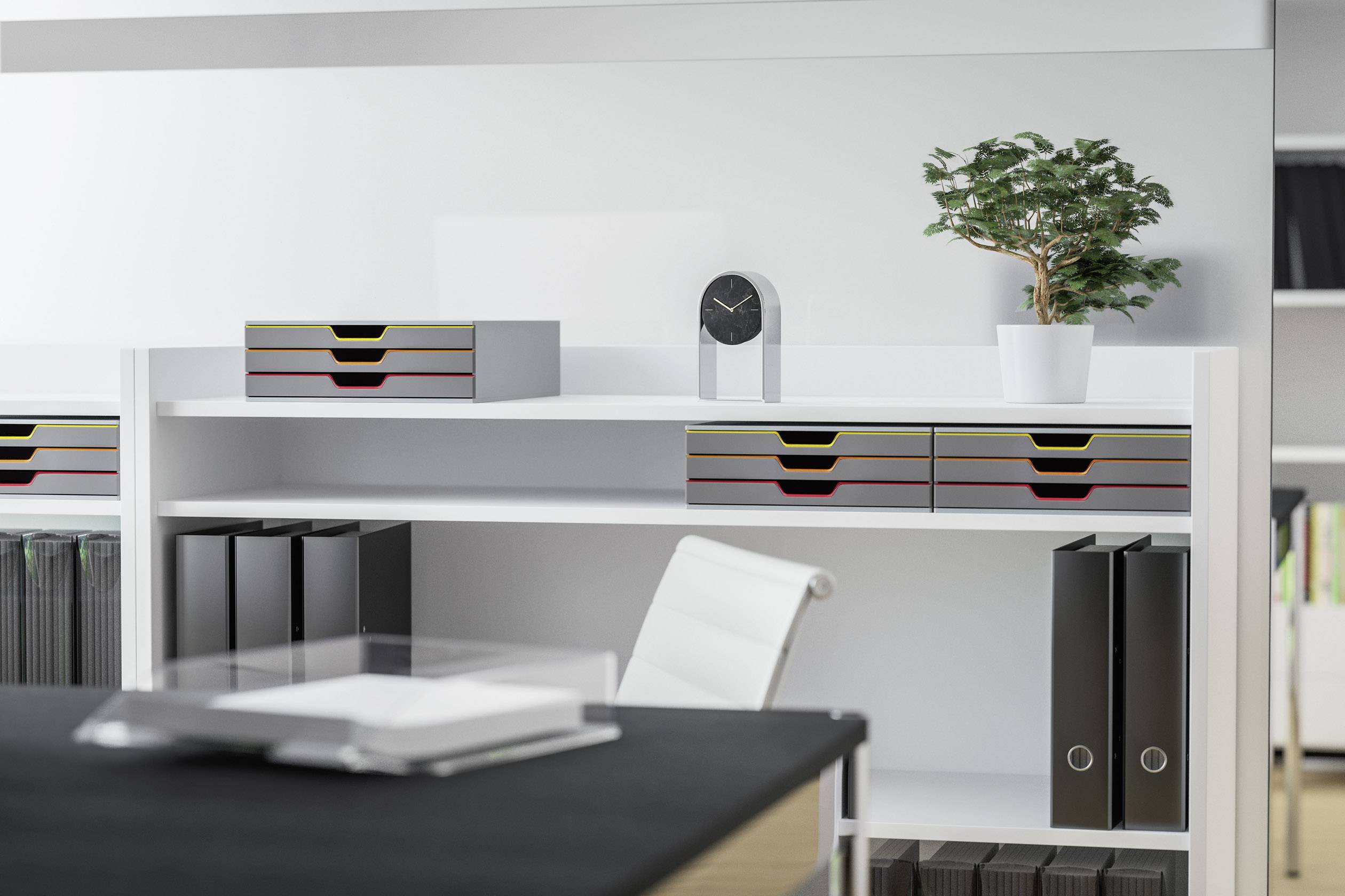 Schreibtisch in modernem Büro mit weißen Regalen, Ordnern, einer kleinen Pflanze und einem minimalistischen schwarzen Wecker.