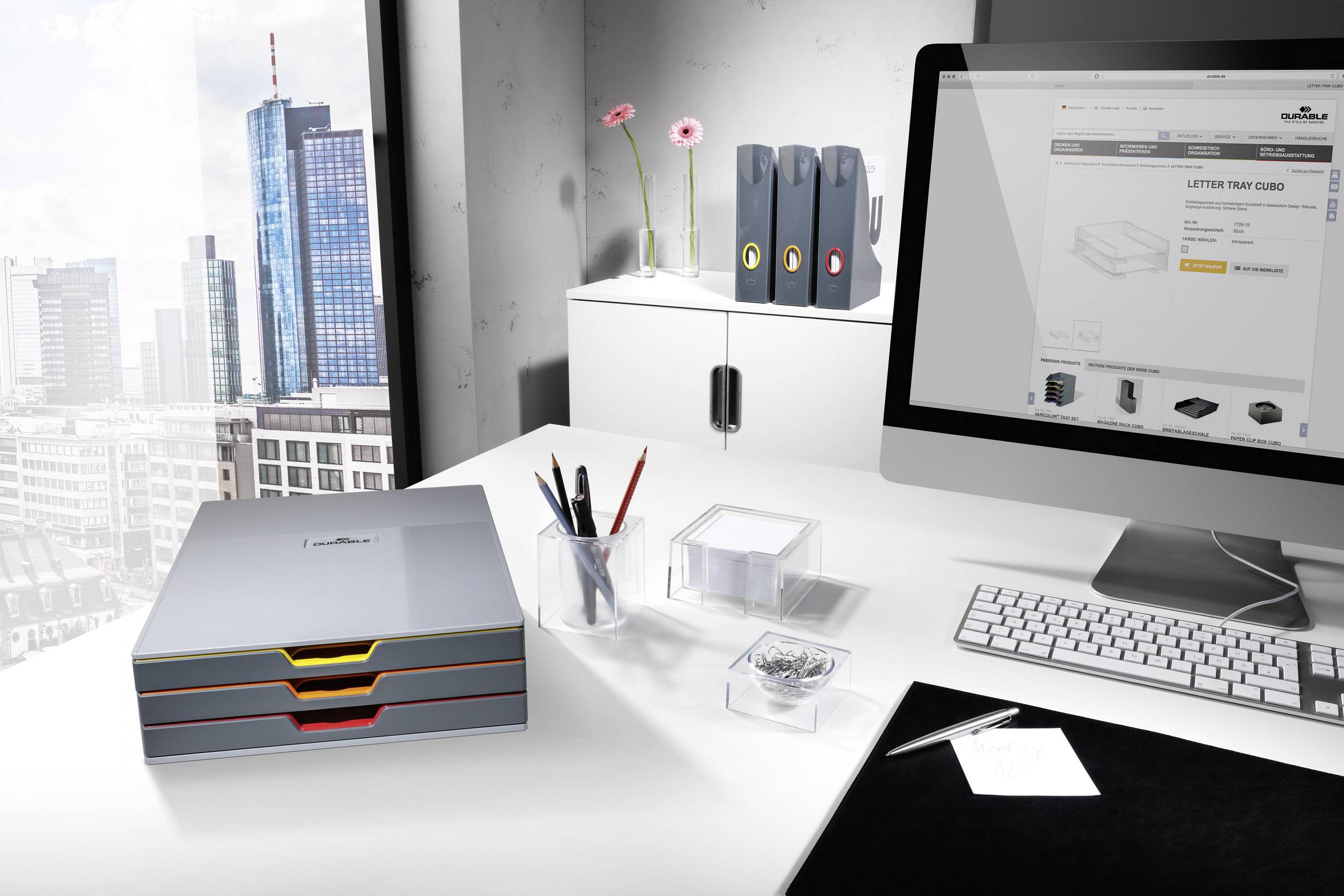 Ein modernes Büro mit Blick auf die Stadt. Auf dem Schreibtisch stehen ein Computer mit Einkaufseite, Ablagen, Stifte und Notizen.