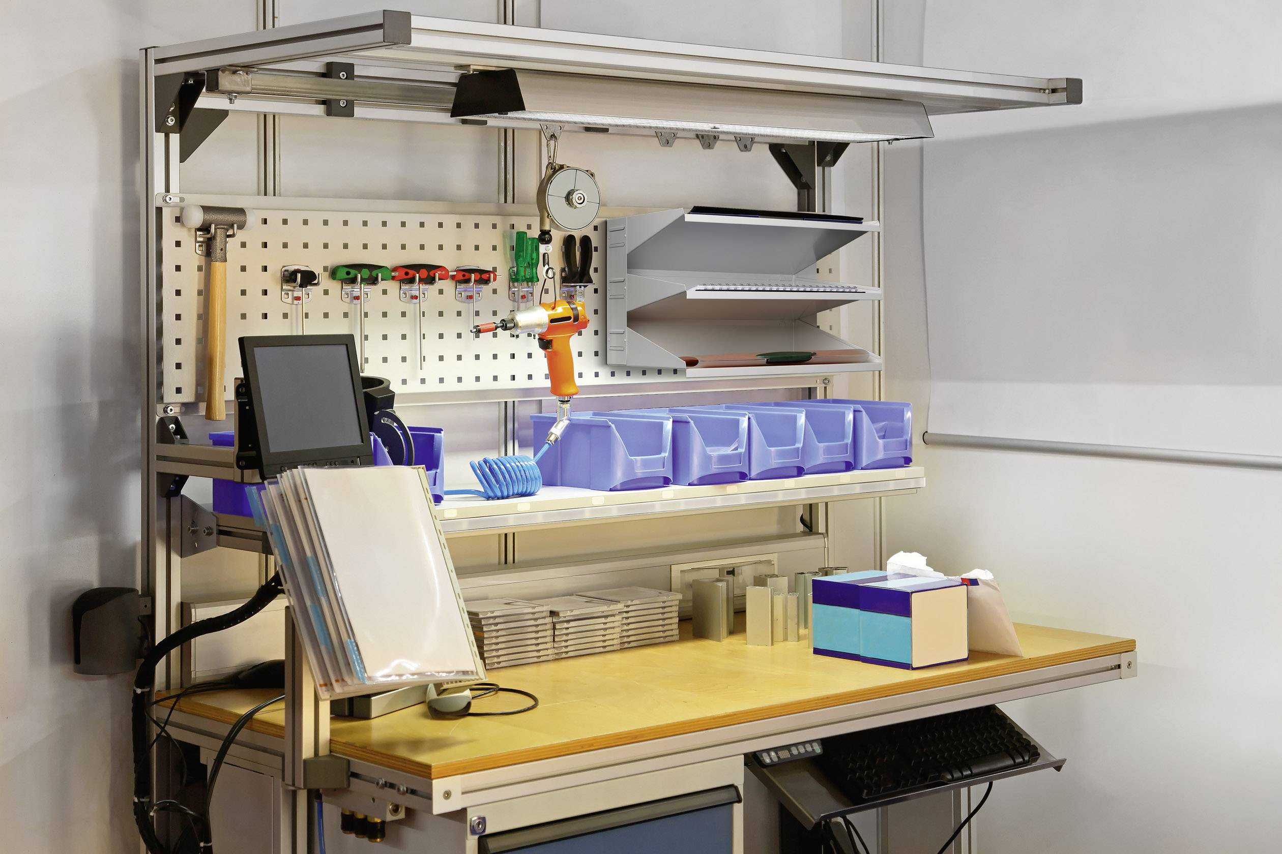 Werkbank mit Werkzeugen an der Wand, Laptop, Dokumentenhalter und sortierten Aufbewahrungsboxen auf dem Tisch.