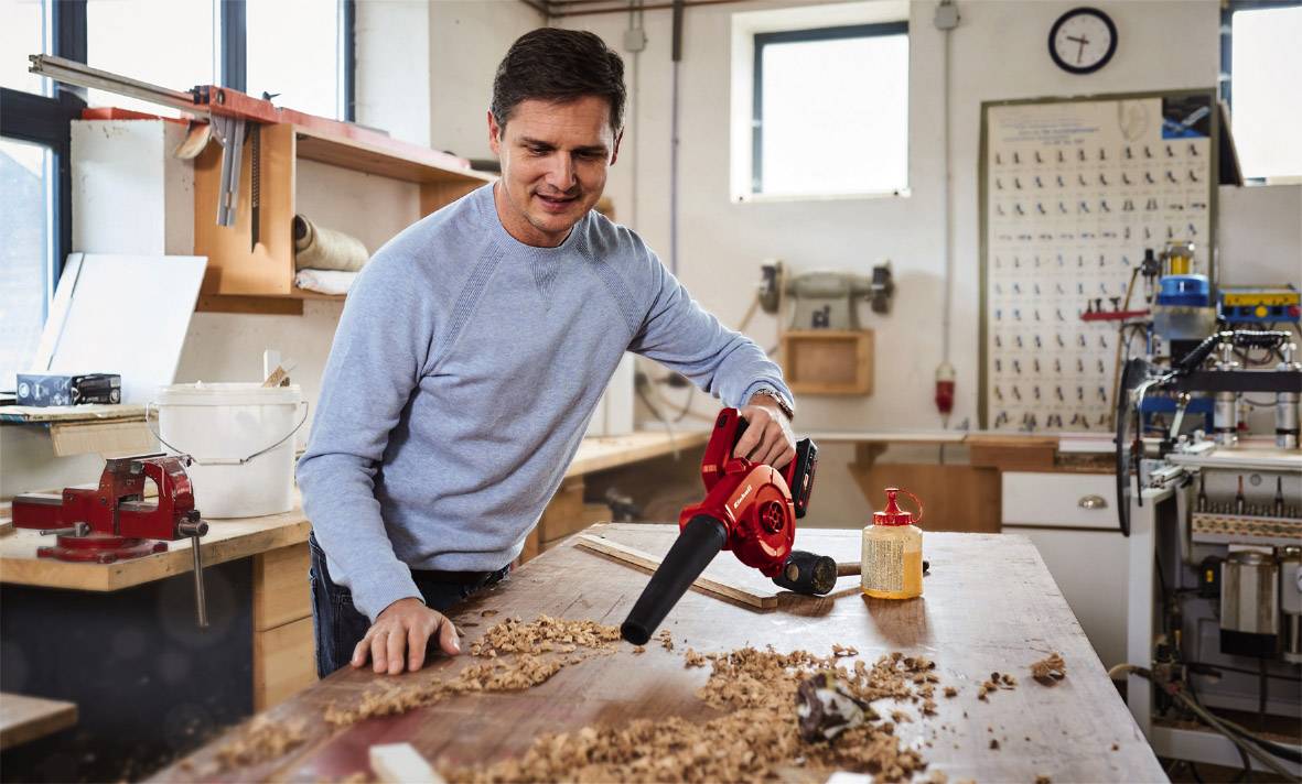 Eine Person in einer Werkstatt benutzt ein rotes Gebläse, um Holzspäne von einem Tisch zu entfernen. Werkzeuge und Schilder im Hintergrund.