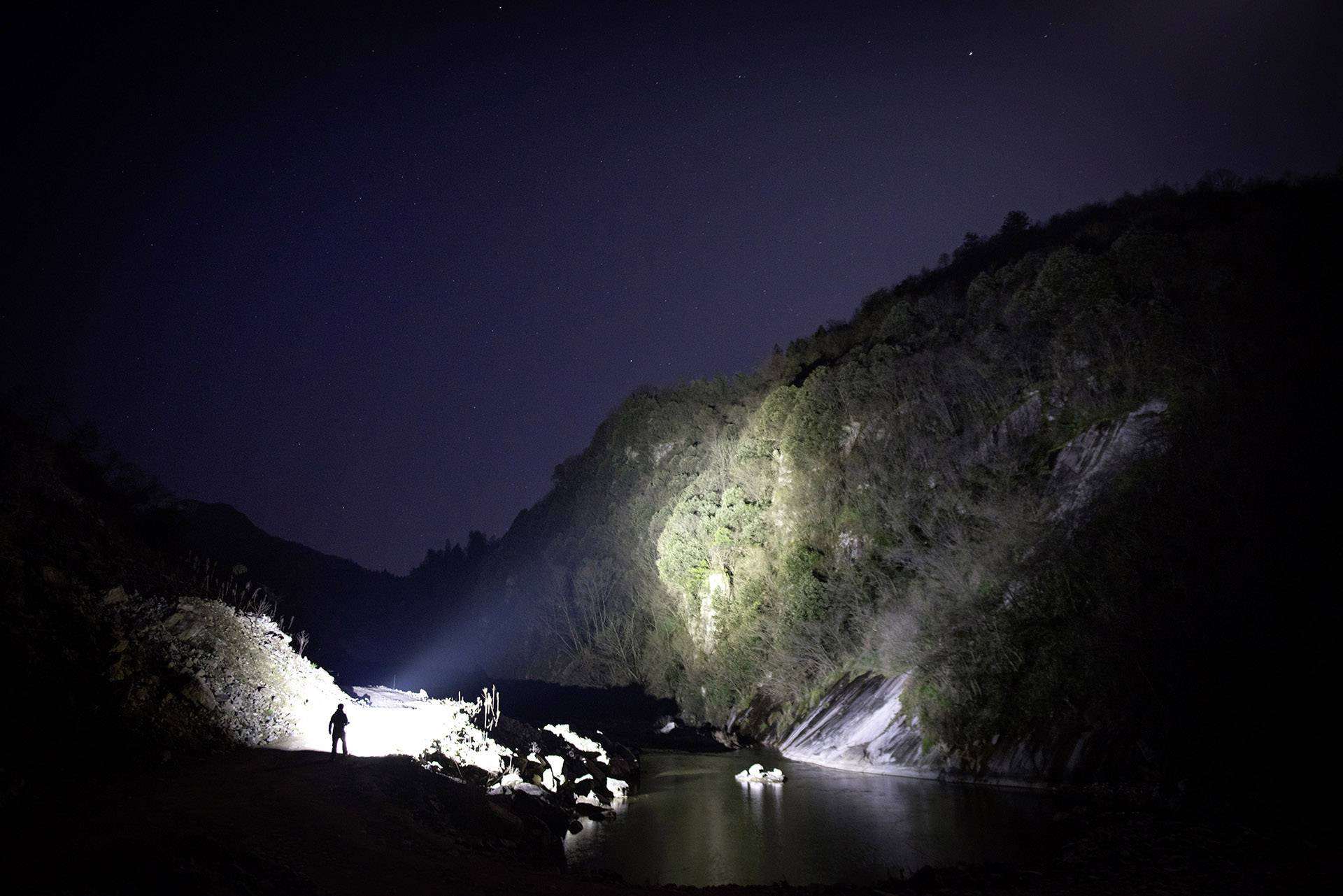 Eine Person steht in dunkler Nacht am Flussufer, eine Taschenlampe beleuchtet die Umgebung. Hügel und Wasser umgeben die Szene.