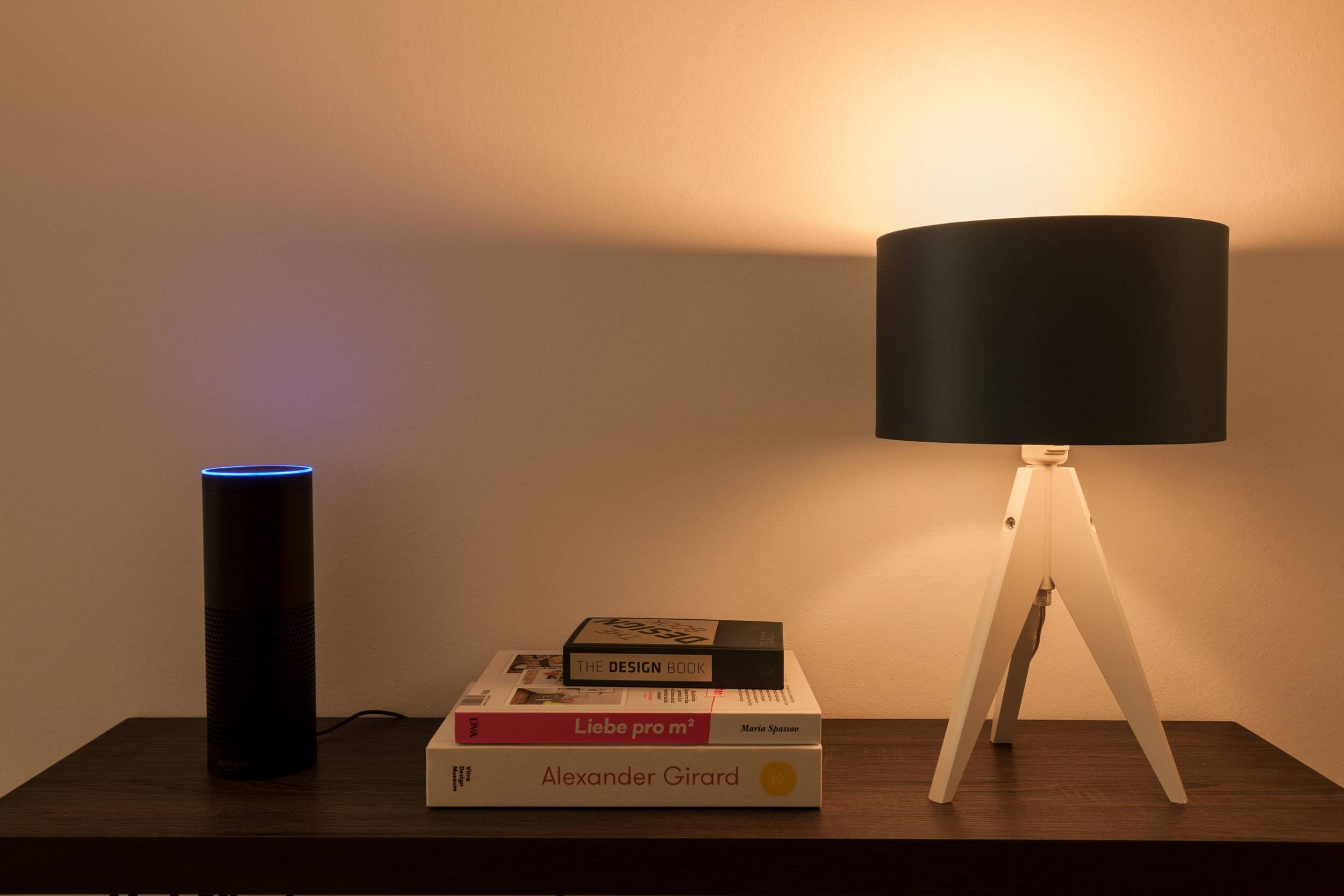 Ein Nachttisch mit einer modernen Lampe und einem Sprachassistenten. Mehrere Designbücher liegen gestapelt daneben.