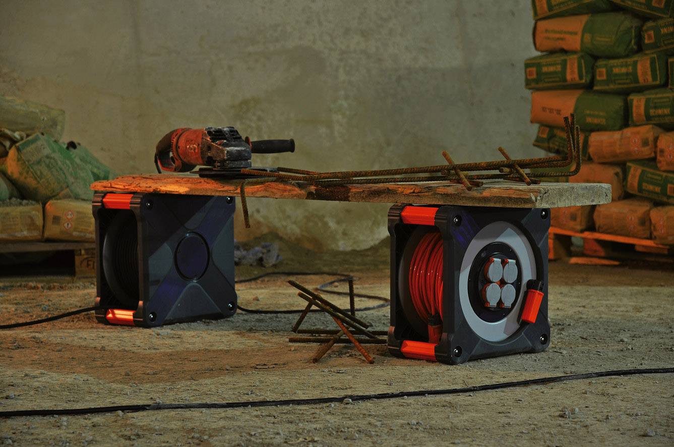 Ein Holztisch auf einer Baustelle, unterstützt von zwei Kabeltrommeln, darauf liegt eine Elektrowerkzeug und Metallstangen. Im Hintergrund sind Säcke gestapelt.