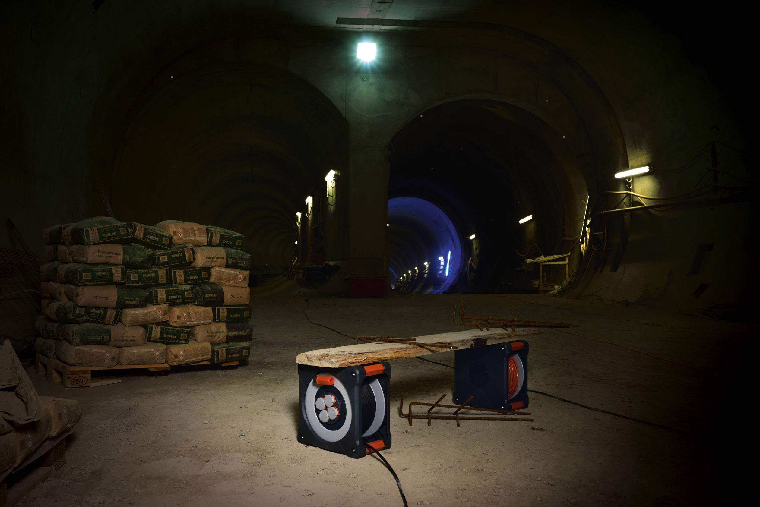 Ein Tunnel im Bau, beleuchtet von Deckenlampen. Im Vordergrund liegen Zementsäcke und ein Ventilator, der auf den Tunnel gerichtet ist.