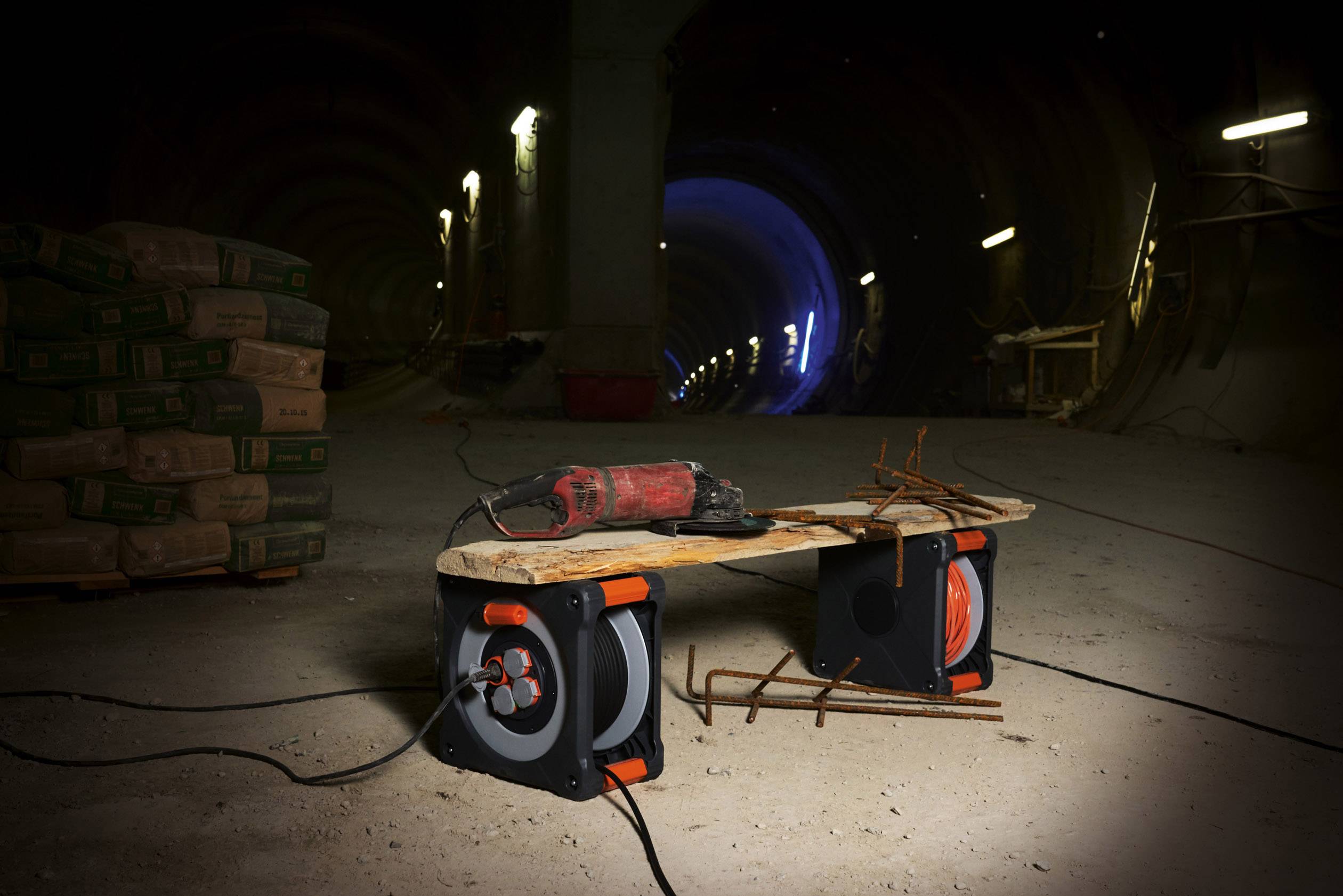 Baustelle in einem Tunnel mit Betonmischern und Bauwerkzeugen im Vordergrund, darunter Kabeltrommeln und eine Bohrmaschine.