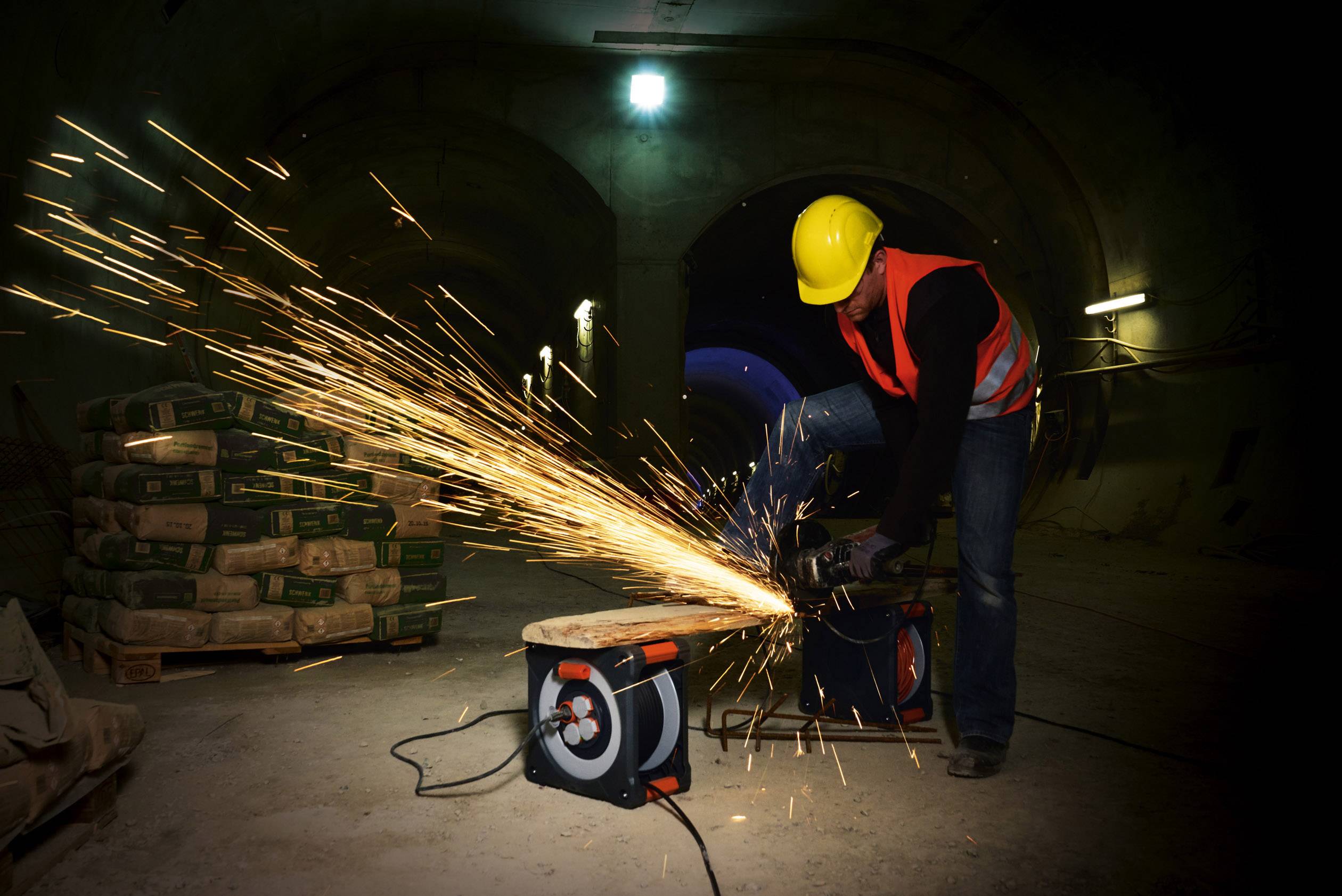 Ein Arbeiter mit gelbem Helm schneidet Metall mit einer Funken sprühenden Schleifmaschine in einem Tunnel.
