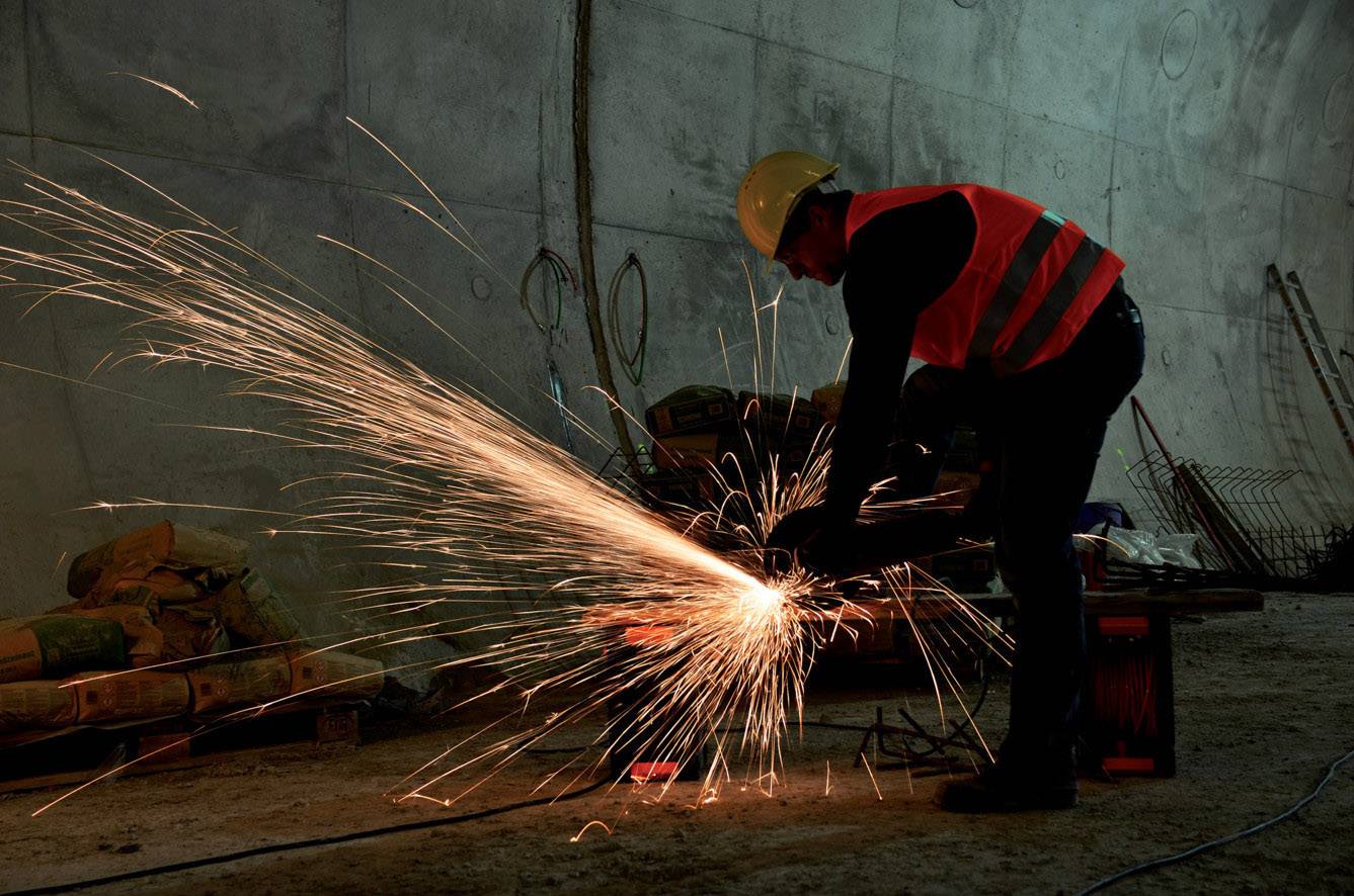 Ein Arbeiter mit Schutzhelm und Warnweste schneidet Metall in einem Tunnel. Funken sprühen hell in einem dunklen, industriellen Umfeld.