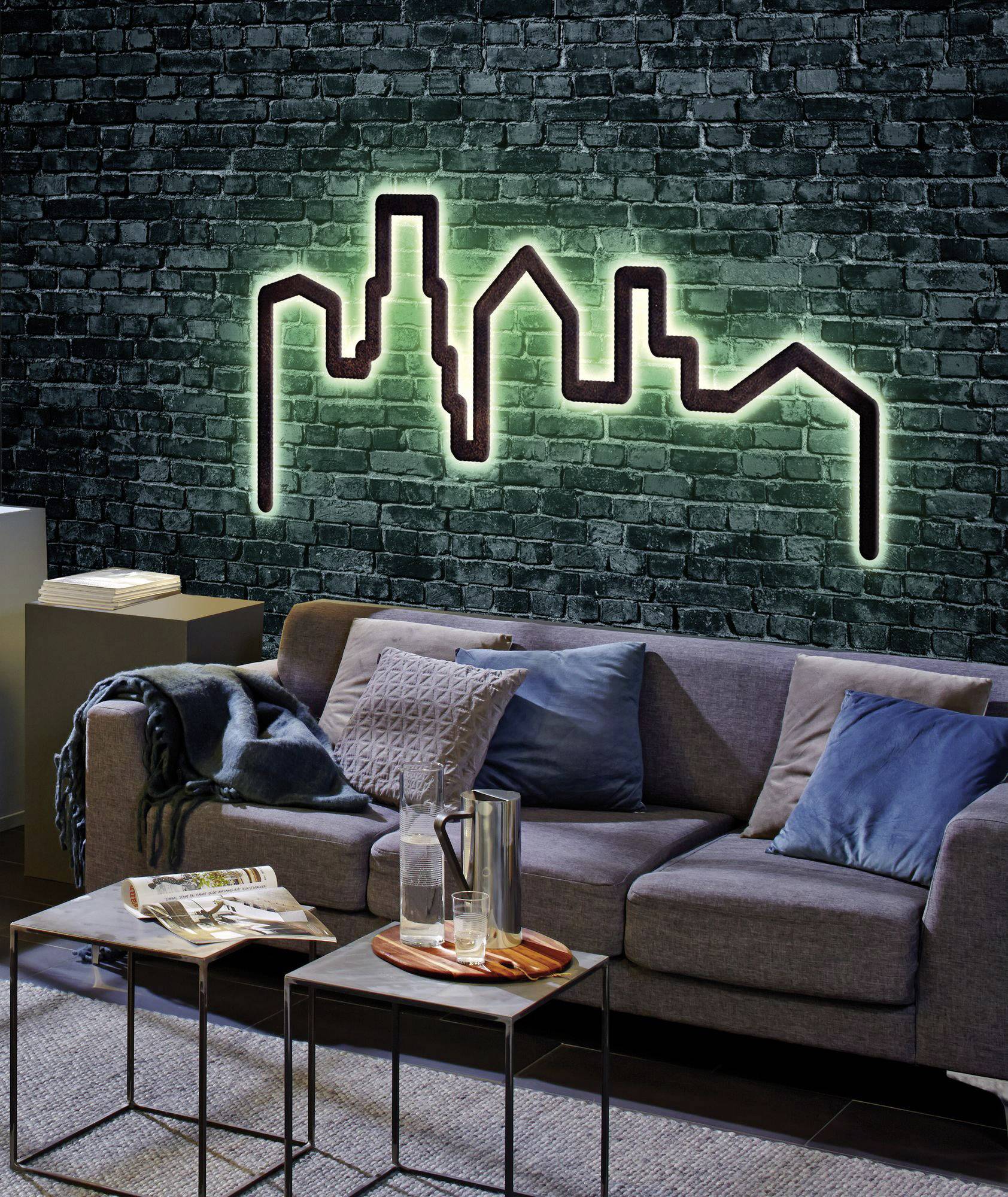 Ein modernes Wohnzimmer mit einem grauen Sofa, blauem Kissen, Decke und zwei kleinen Tischen. An der Wand eine leuchtende Skyline-Dekoration.