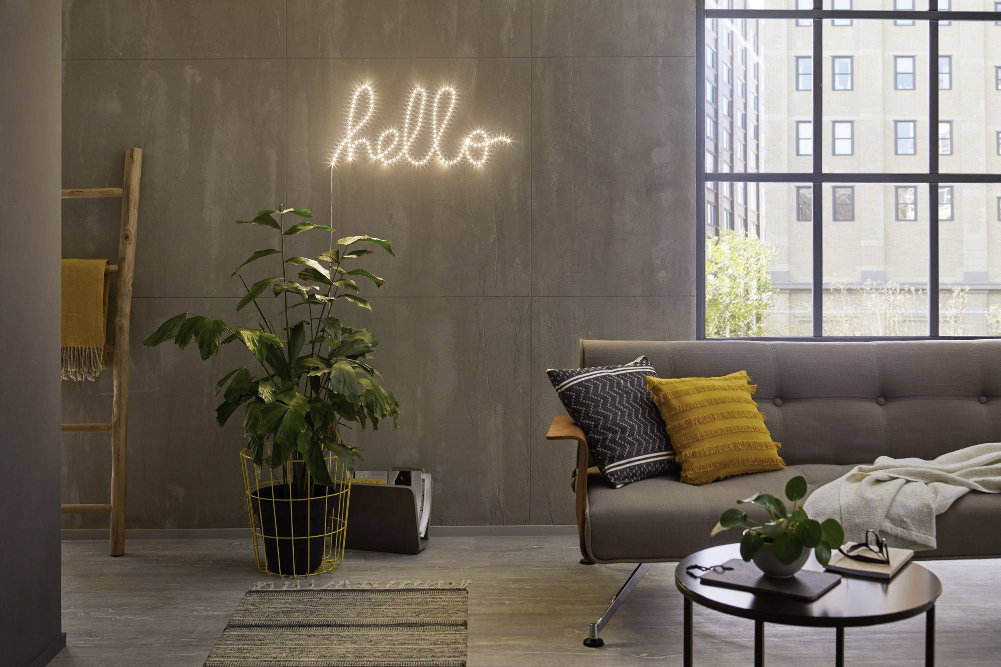 Ein Wohnzimmer mit einem modernen Sofa, Pflanzen und einem 'hello'-Leuchtschild an der Wand. Große Fenster lassen Tageslicht herein.