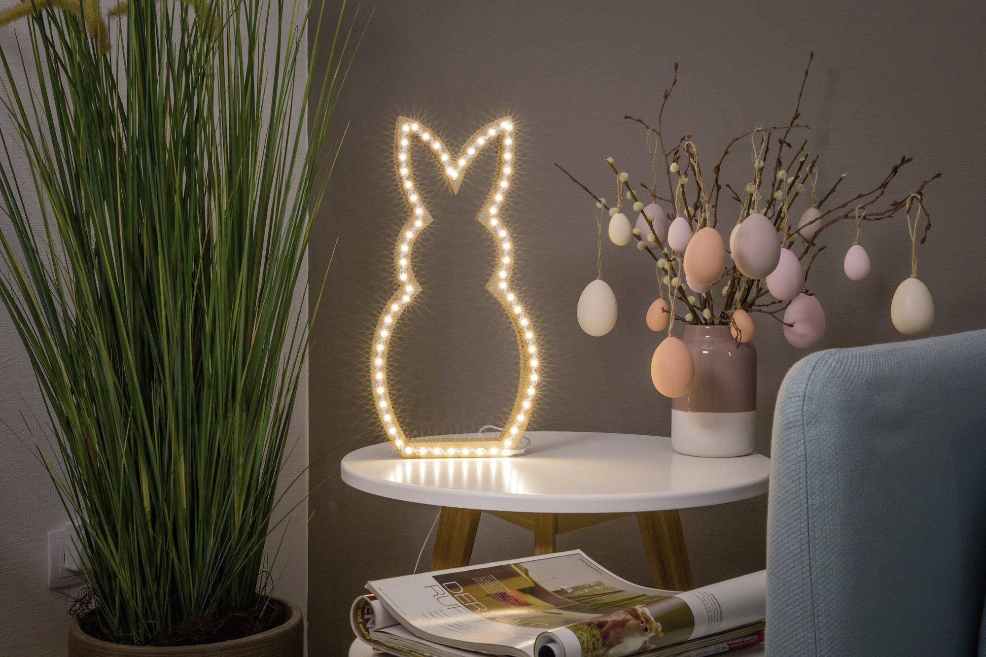 Ein beleuchteter Hasenrahmen steht auf einem weißen Tisch neben einem Strauß mit hängenden Ostereiern. Links steht eine Pflanze.