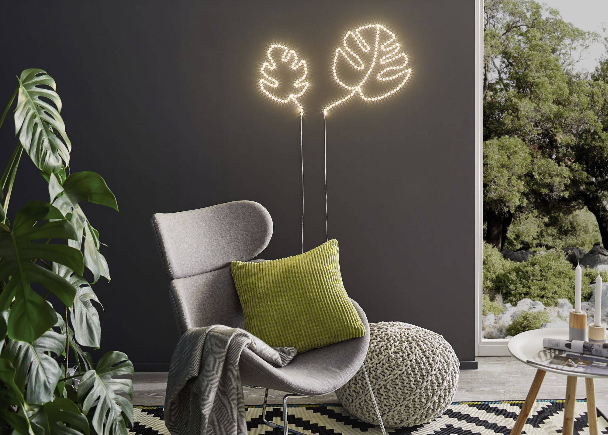 Ein moderner, grauer Sessel mit grünem Kissen steht vor einer Wand mit leuchtenden Pflanzenmotiven. Daneben ein großes Fenster mit Gartenblick.