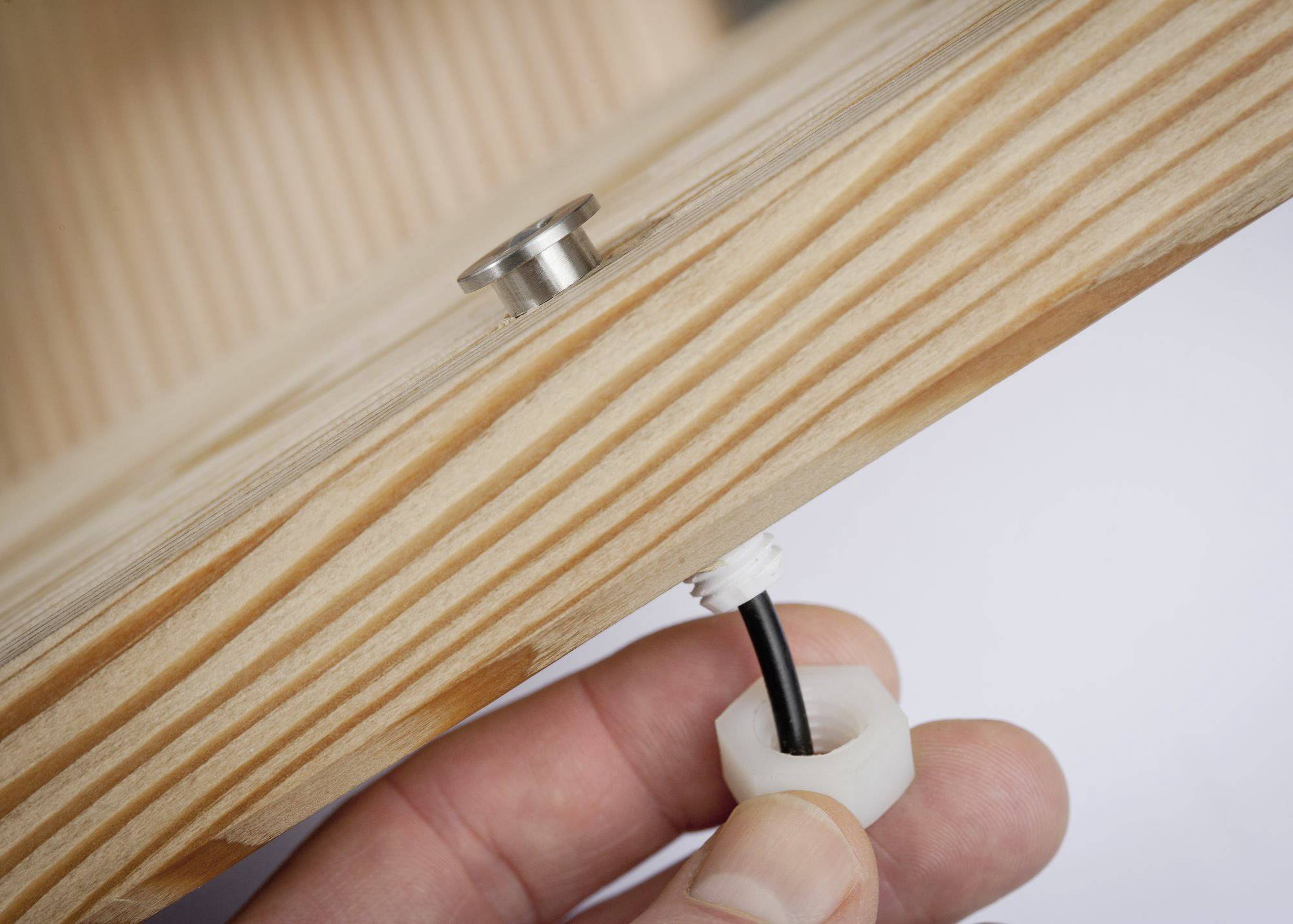 Hand zieht schwarzes Kabel durch eine Holzkonstruktion mit Metallbolzen und weißer Kunststoffmutter, Nahaufnahme.