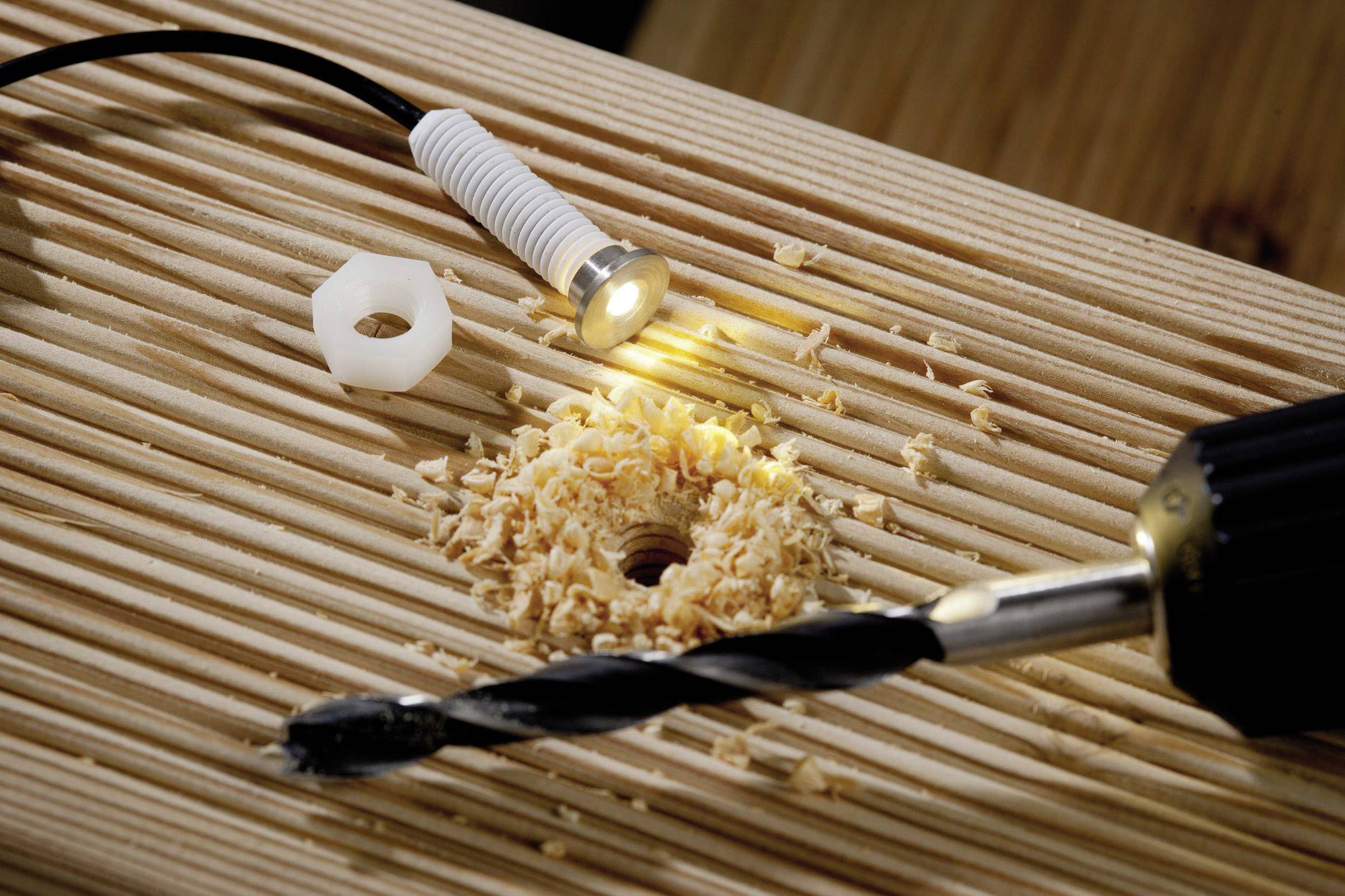 Nahaufnahme eines Holzstücks, das mit einem Bohrer ein Loch erhält. Holzspäne sind um das Loch verteilt, eine kleine Lampe leuchtet.