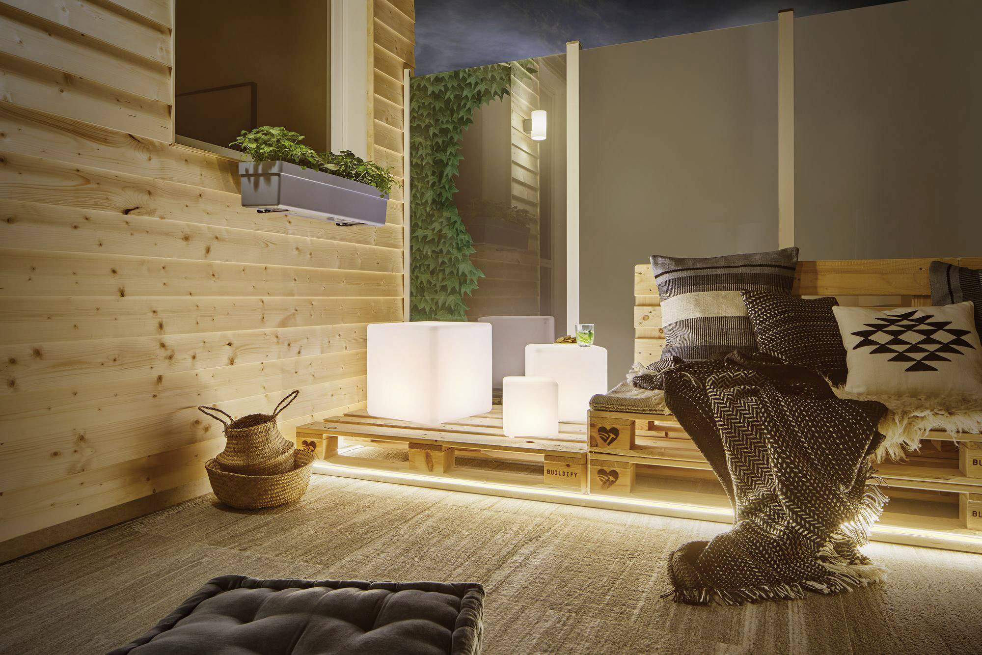 Gemütliche Terrasse bei Nacht, mit Holzpalettenmöbeln, kuscheligen Decken und indirekter Beleuchtung. Pflanzen im Hintergrund.