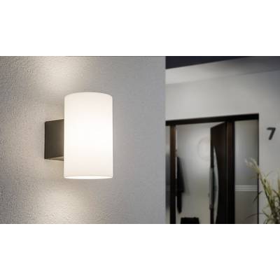 Eine moderne Wandleuchte mit zylindrischem, weißem Schirm leuchtet in einem Flur. Im Hintergrund ist eine leicht geöffnete Tür sichtbar.