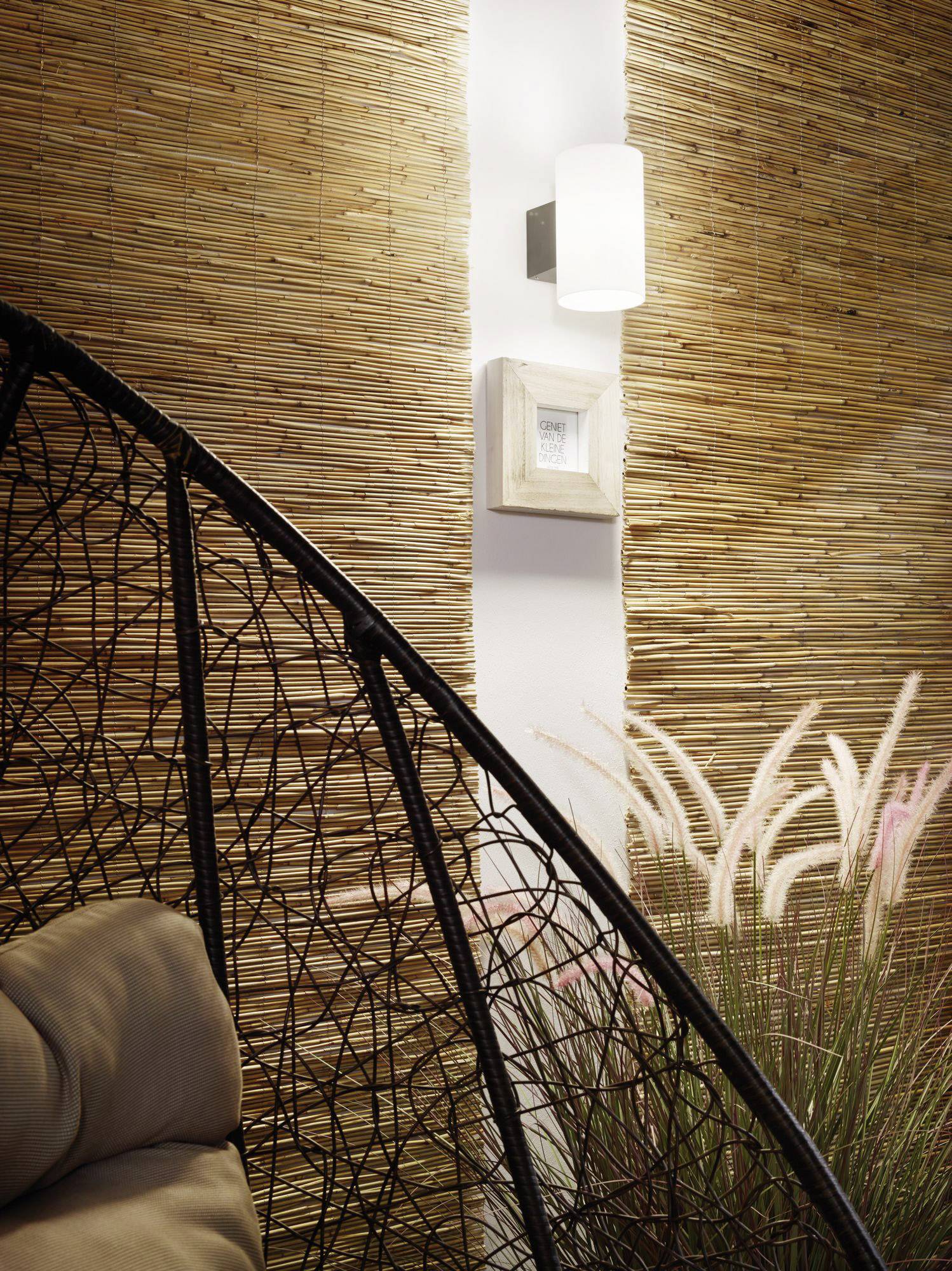 Korbschaukel im Vordergrund vor einer Wand aus Bambus oder Holz, mit einem Wandlicht und dekorativen Gräsern daneben.