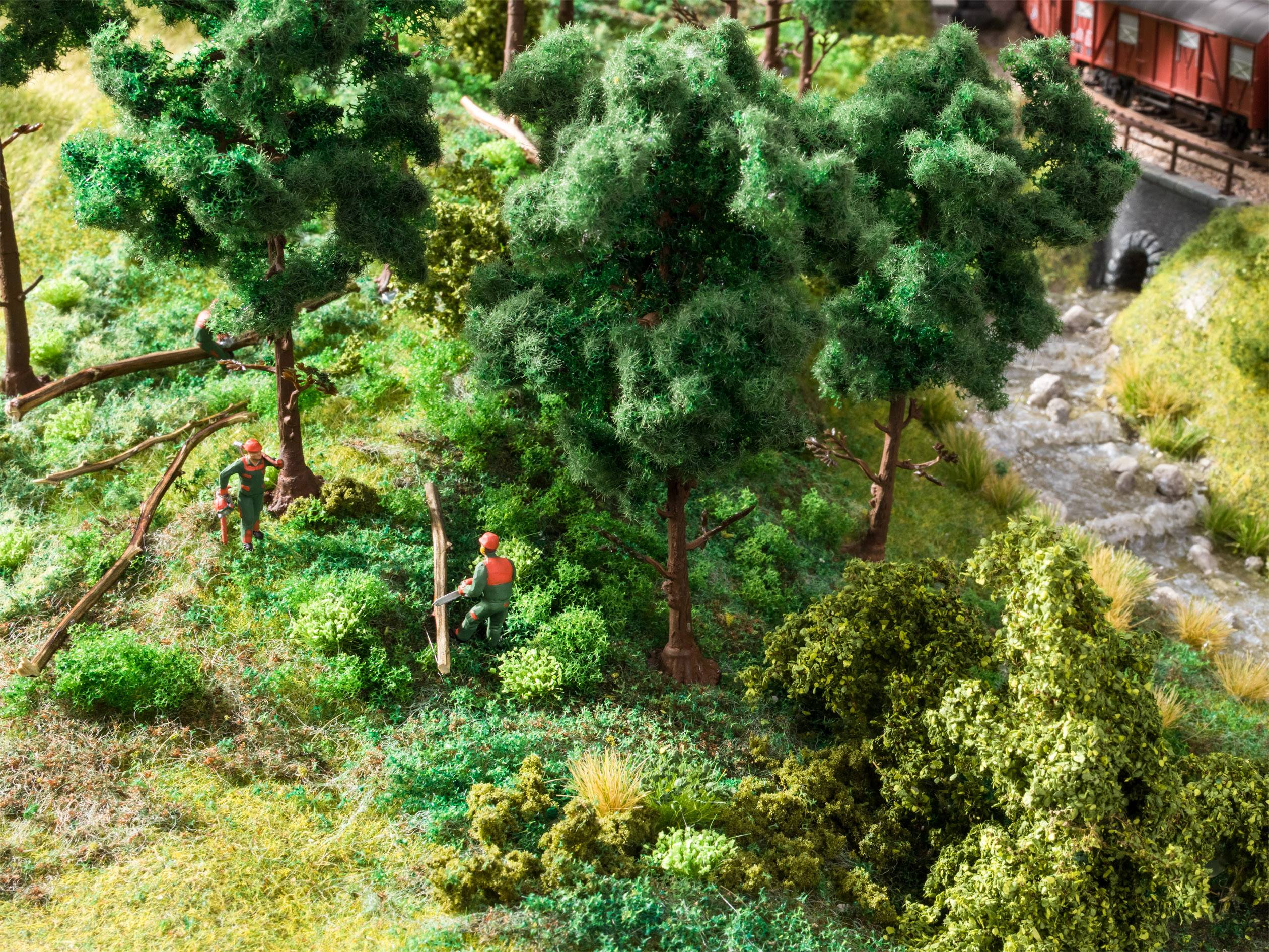 Zwei Miniaturfiguren mit Helmen fällen einen Baum in einem Wald, nahe eines kleinen Flusses mit Brücke im Hintergrund.