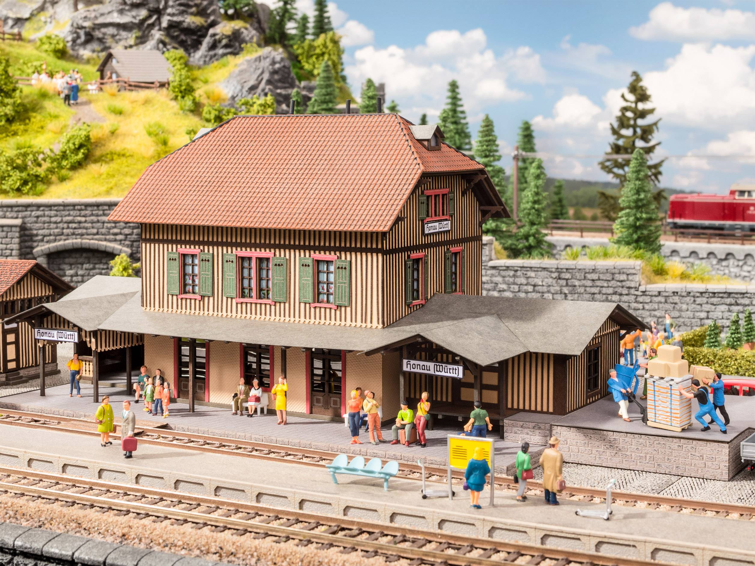 Modelleisenbahn-Szene mit einem Bahnhof, umgeben von Miniaturmenschen, die warten oder sich unterhalten. Im Hintergrund sind Bäume und ein Zug.