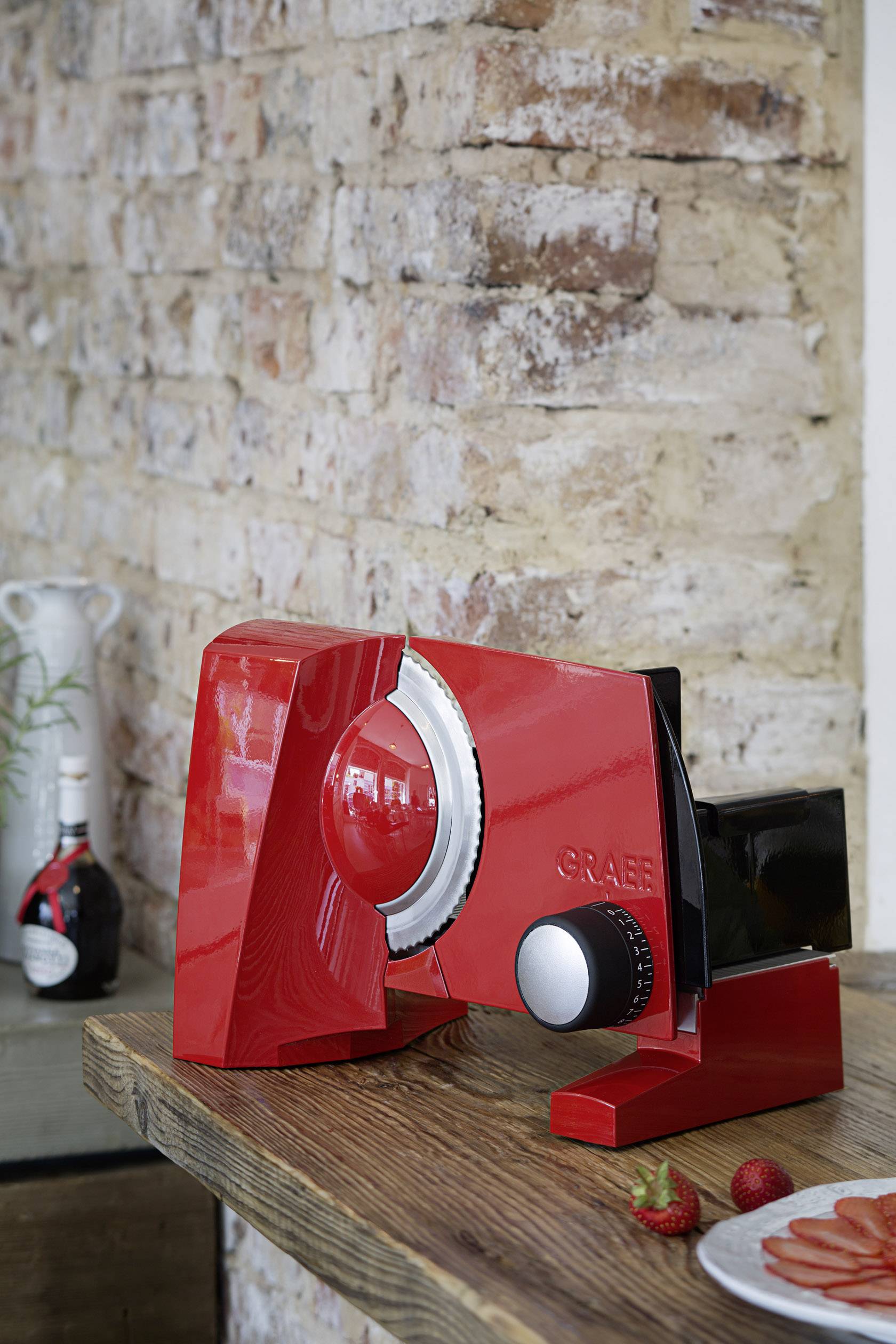 Roter Allesschneider auf Holztisch vor Backsteinwand, neben Erdbeeren und aufgeschnittener Salami. Im Hintergrund Flasche mit italienischem Design.
