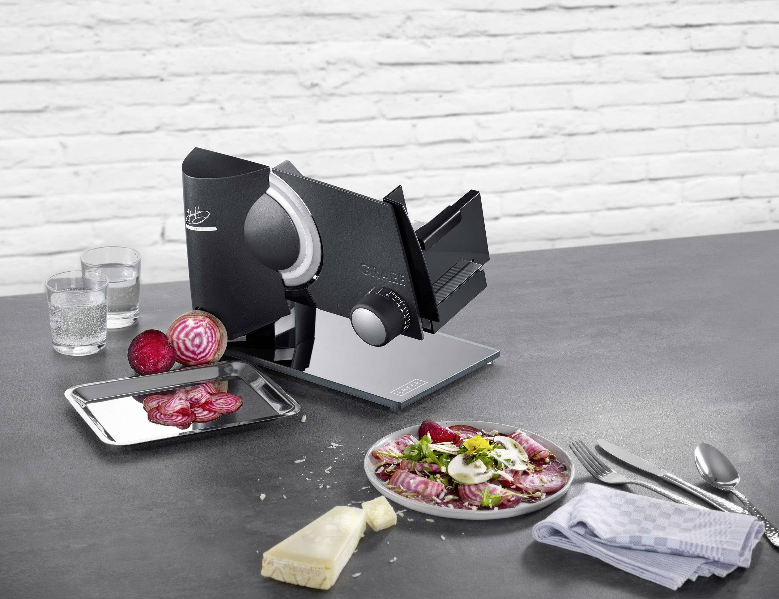 Elektrische Schneidemaschine auf Tisch mit aufgeschnittenem Gemüse, Käse und Besteck, im Hintergrund weiße Ziegelwand.