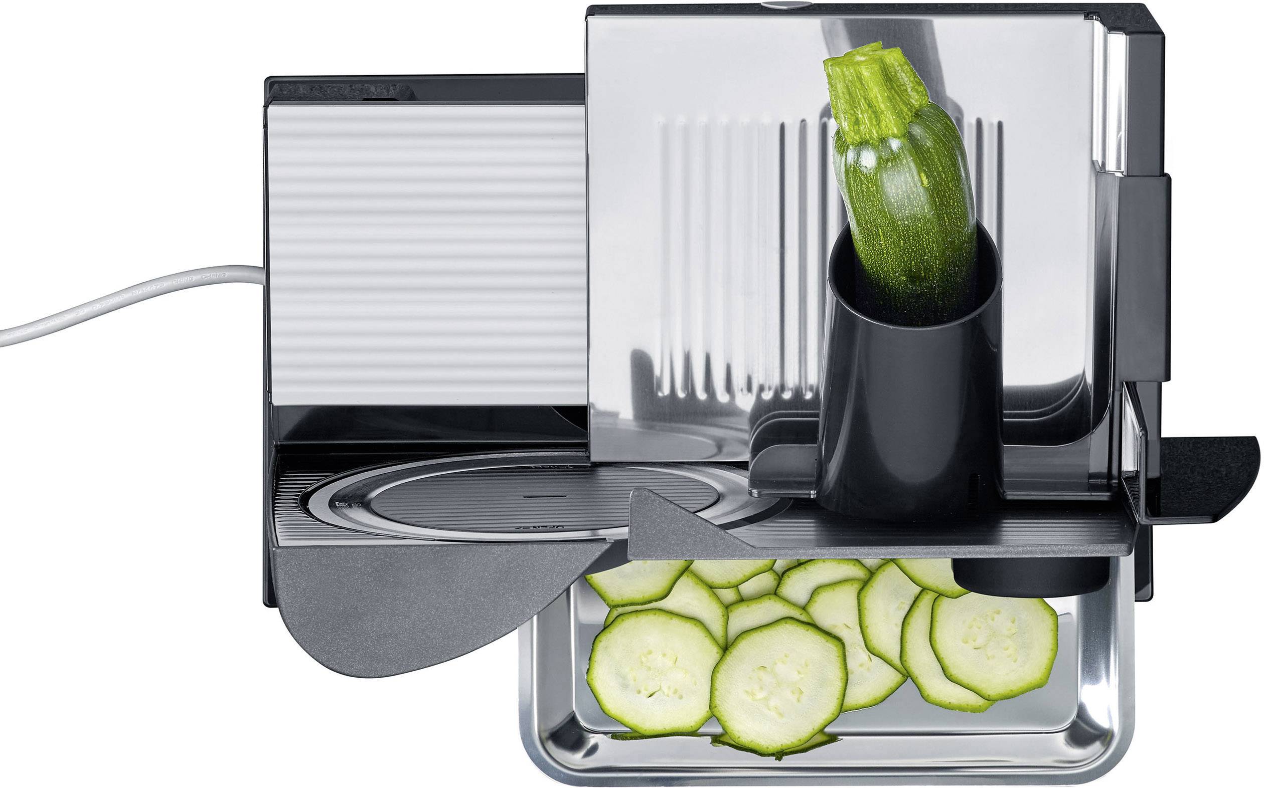 Eine elektrische Aufschnittmaschine schneidet eine grüne Zucchini in gleichmäßige Scheiben, die in einem Behälter gesammelt werden.