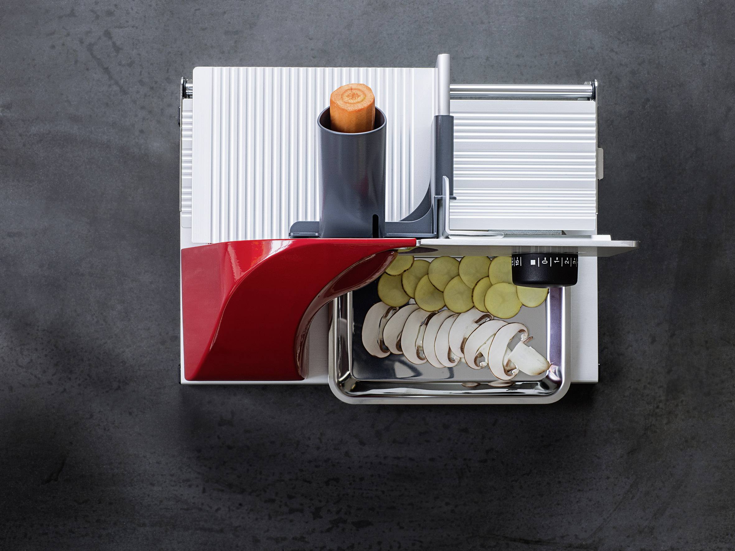 Gemüseschneider mit Karotte oben und Scheiben von Zucchini und Pilzen darunter auf schwarzer Oberfläche.