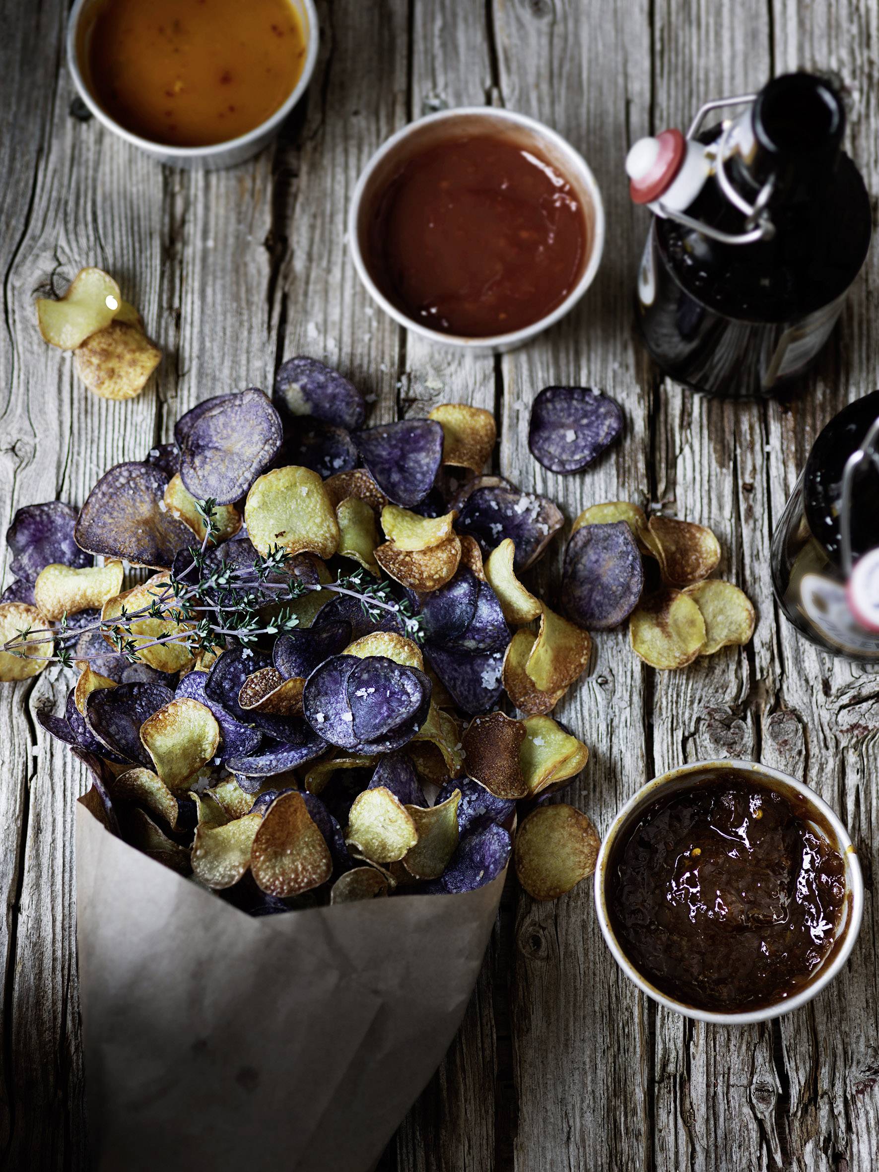 Eine Tüte mit hausgemachten Chips aus violetten und goldenen Kartoffeln, serviert mit verschiedenen Dips und Getränken auf einem Holztisch.