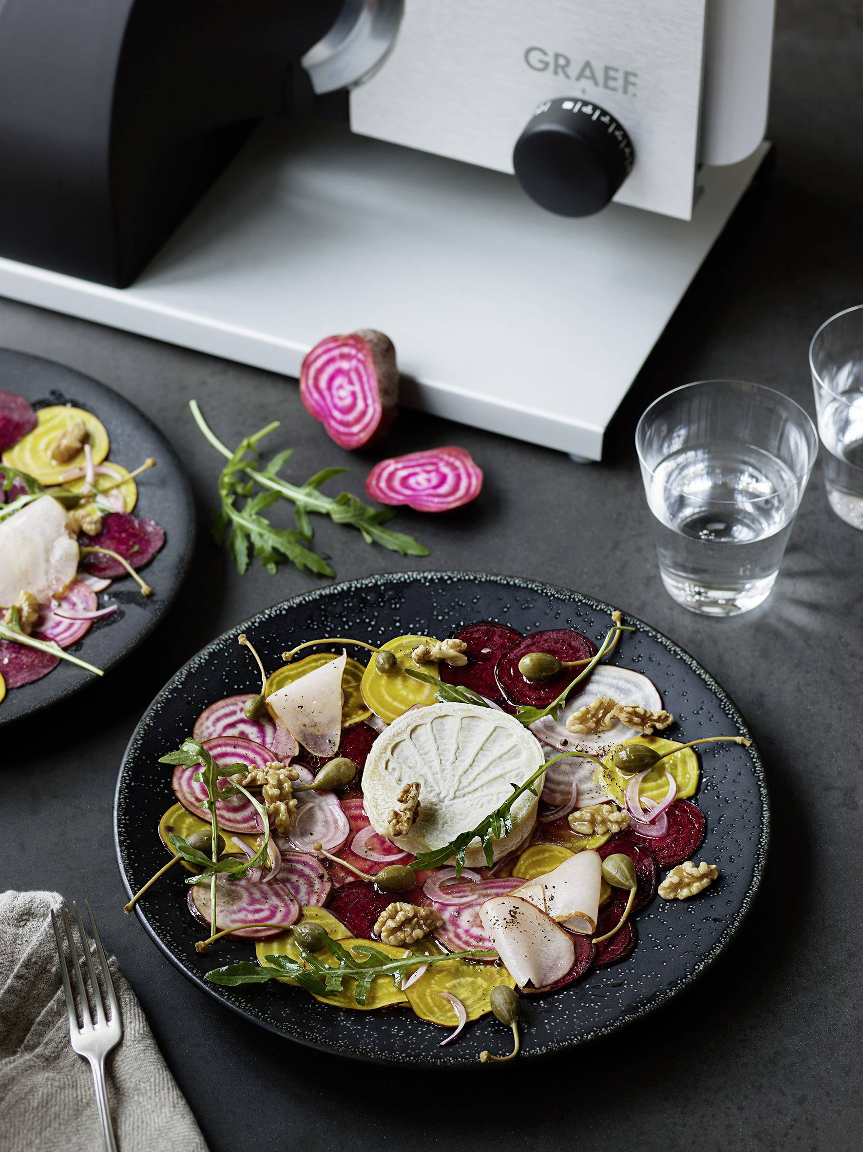 Ein Teller mit gemischtem Salat aus Roter Bete, Radieschen, Walnüssen und Käse, mit zwei Gläsern Wasser und einer Schneidemaschine im Hintergrund.