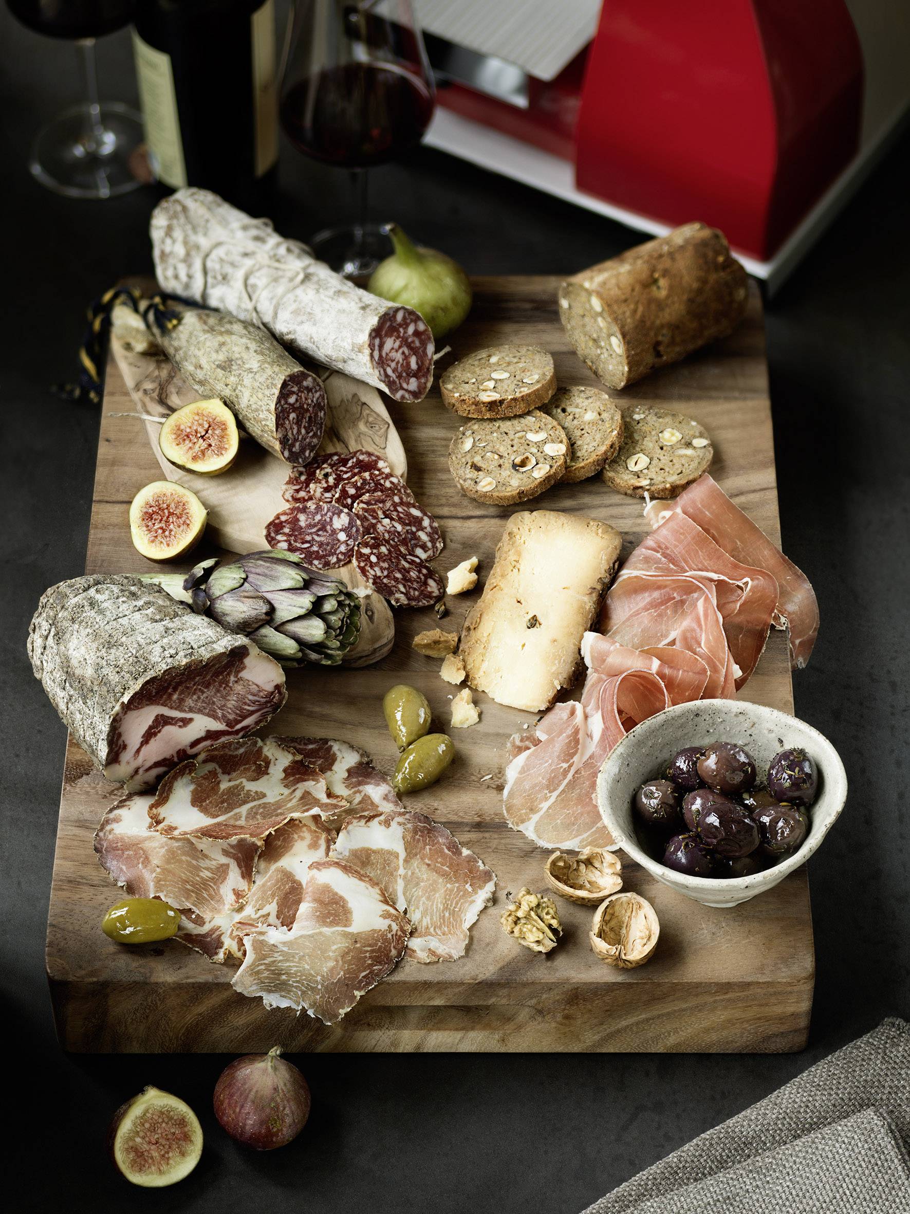 Ein hölzernes Servierbrett mit verschiedenen Wurstsorten, Käse, Oliven, Feigen und Crackern. Zwei Gläser Rotwein im Hintergrund.