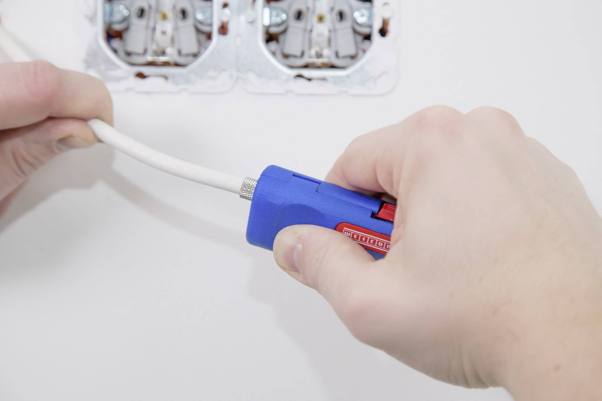 Eine Person benutzt ein Abisolierwerkzeug, um ein Kabel freizulegen. Im Hintergrund sind zwei Steckdosen sichtbar.