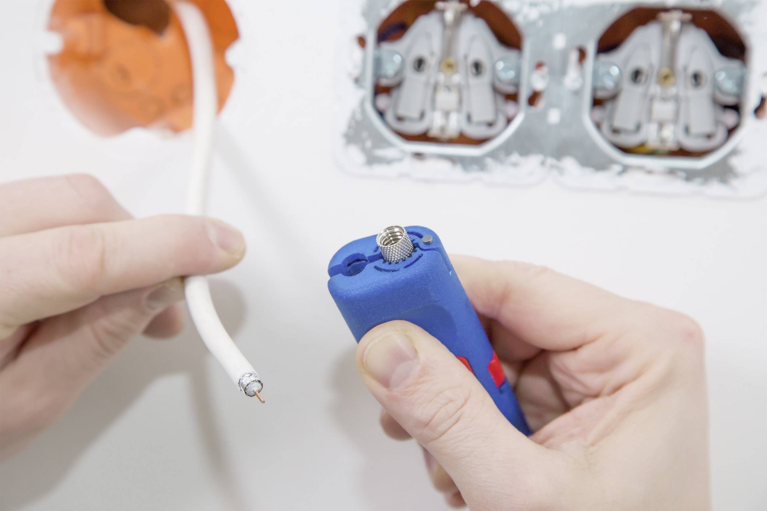 Eine Person hält ein Werkzeug und ein Kabel neben einer Steckdose. Das Werkzeug wird verwendet, um die Isolierung des Kabels zu entfernen.