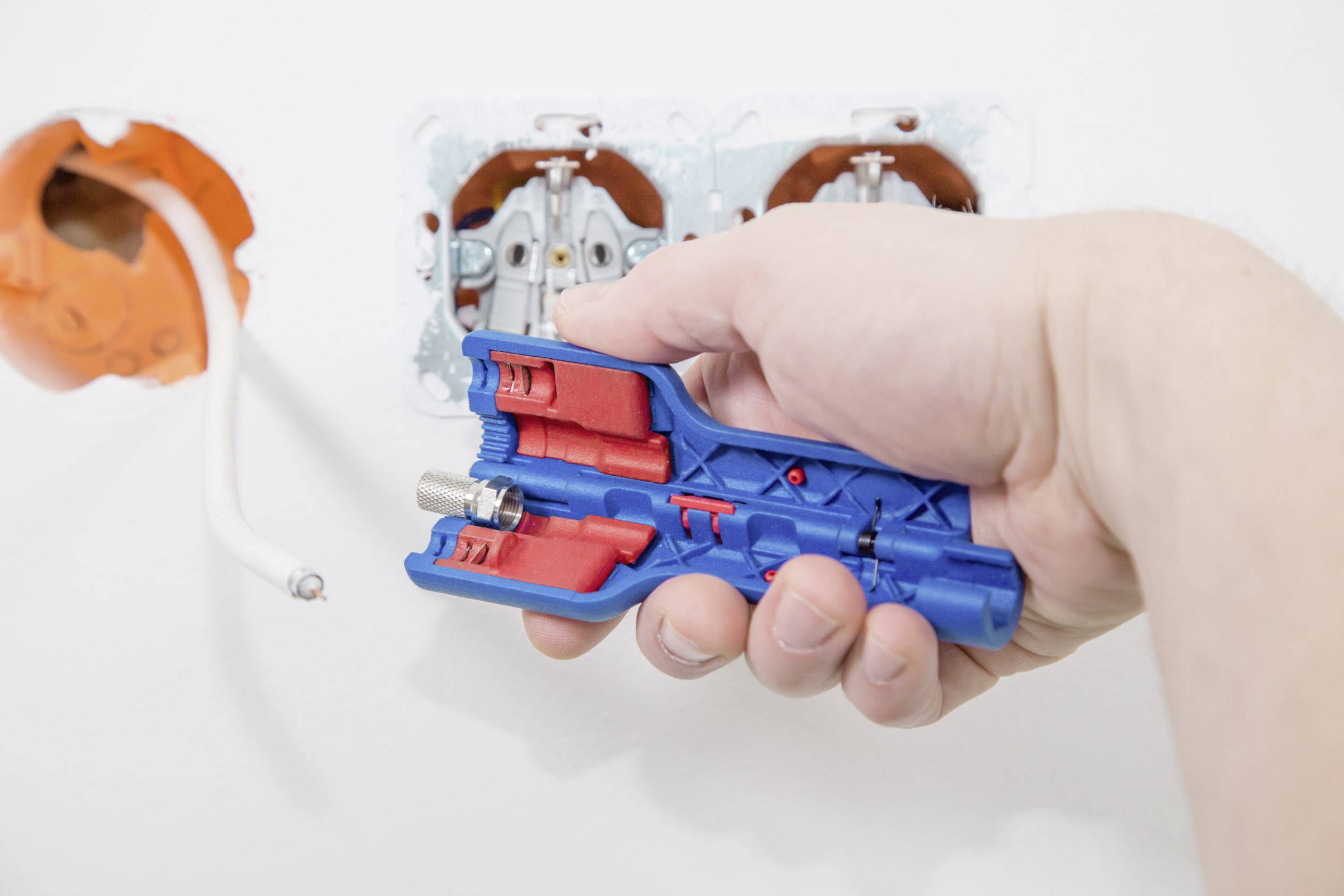 Eine Hand hält ein blau-rotes Werkzeug vor einer ungeöffneten Steckdose. Ein Kabel hängt lose aus einem orangefarbenen Anschluss.