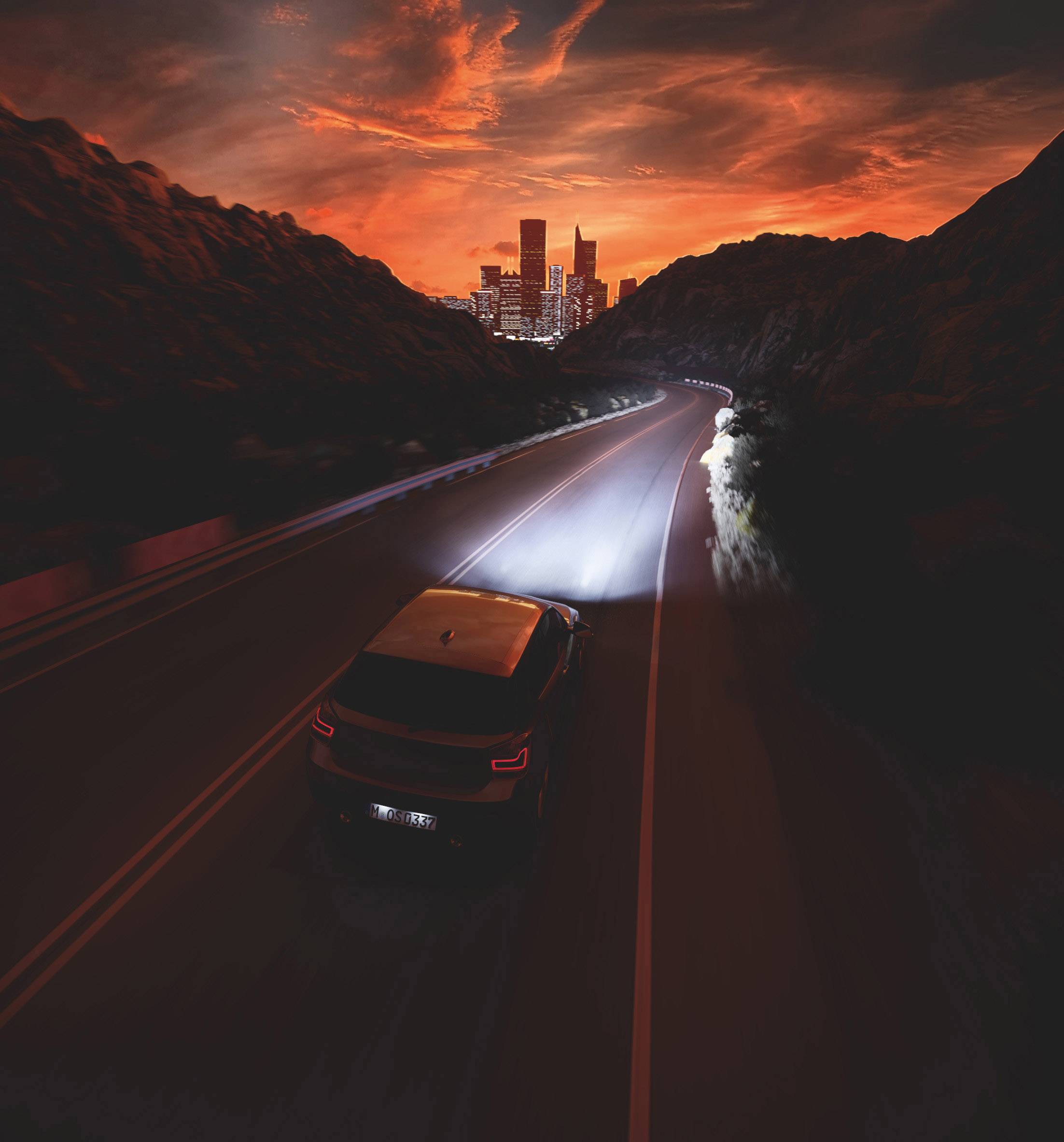 Ein Auto fährt nachts auf einer kurvigen Straße in Richtung einer beleuchteten Stadt mit roten, dramatischen Wolken am Himmel.