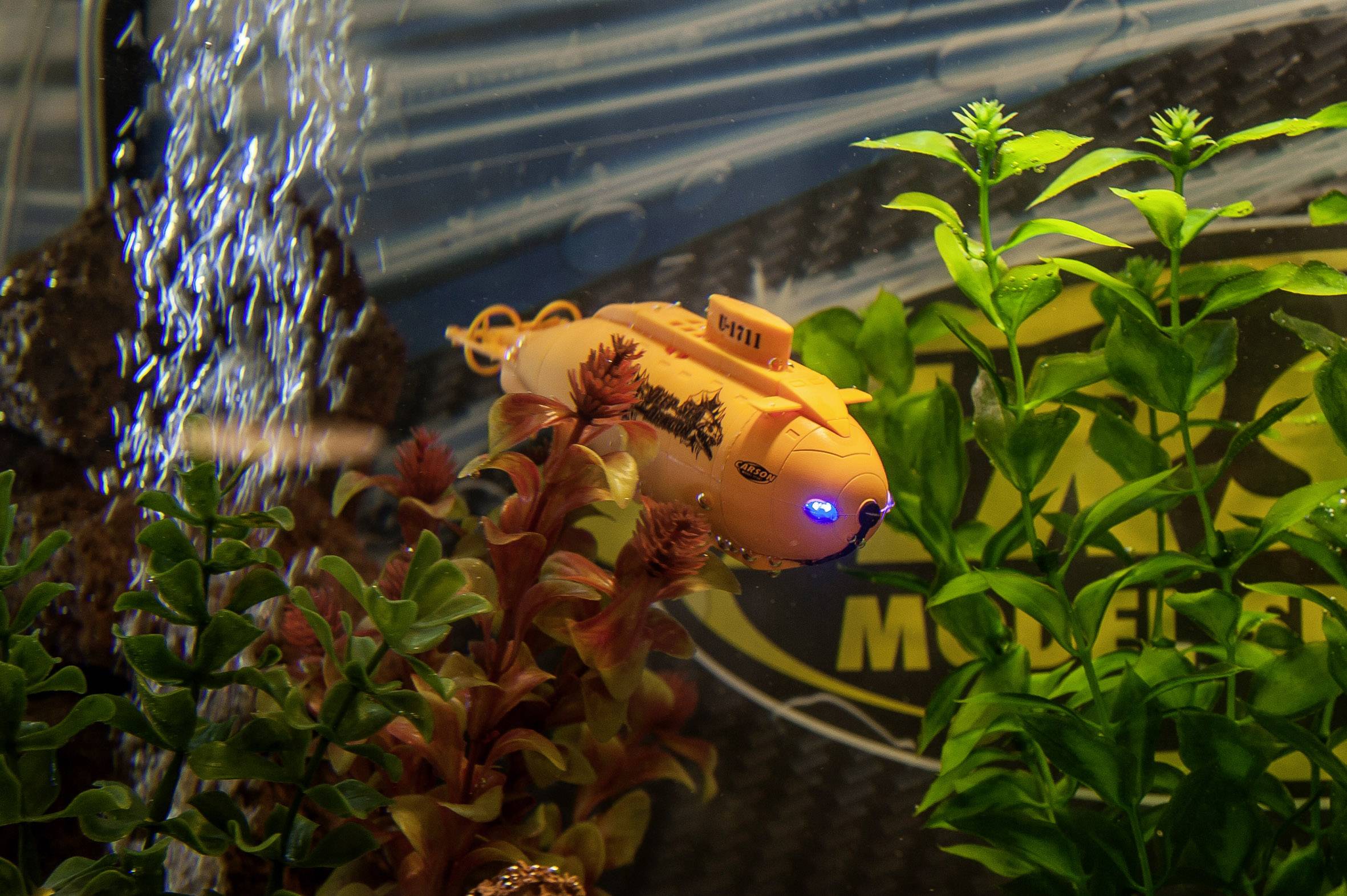 Ein kleines, oranges Spielzeug-U-Boot mit blauem Licht taucht in einem Aquarium zwischen grünen Pflanzen und Luftblasen.
