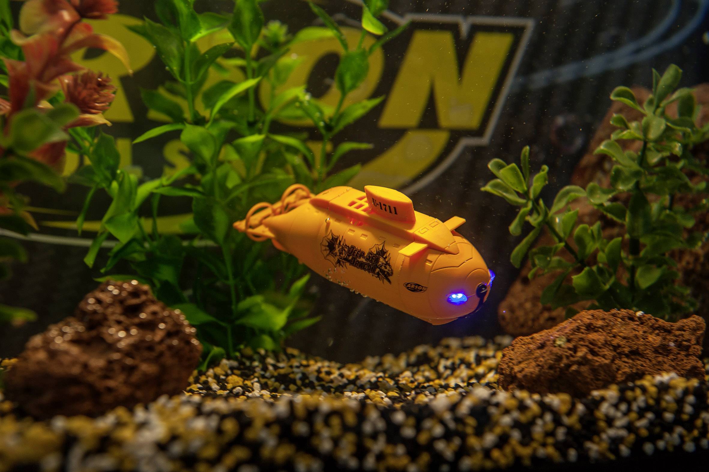 Ein gelbes Mini-U-Boot schwimmt in einem Aquarium mit Pflanzen und Steinen, beleuchtet von einem blauen Licht an der Vorderseite.