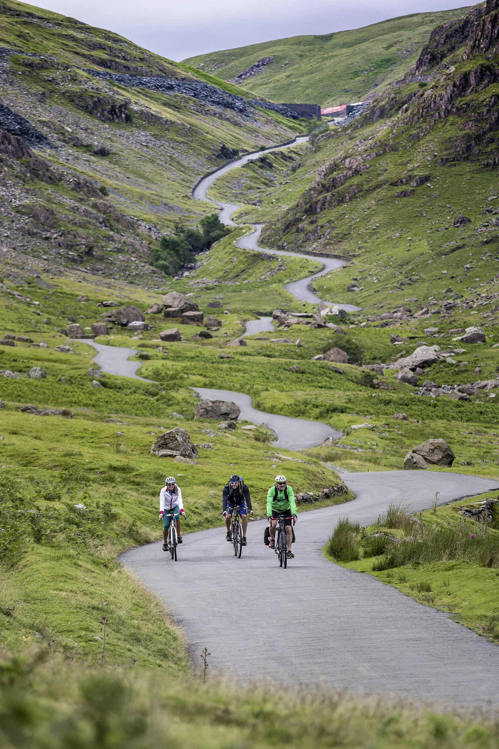 Drei Radfahrer fahren einen kurvigen Weg in einer grünen Hügellandschaft hinab. Der Himmel ist bewölkt und eine Hütte ist in der Ferne sichtbar.