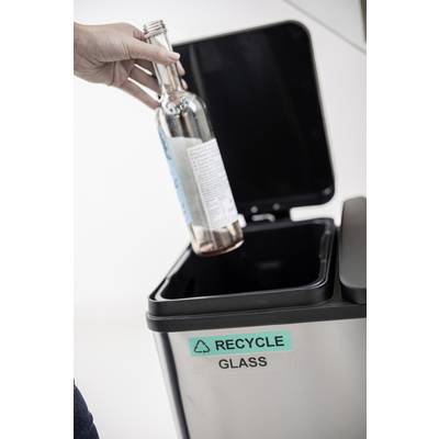 Eine Person wirft eine leere Glasflasche in einen Mülleimer mit der Aufschrift 'Recycle Glass'.