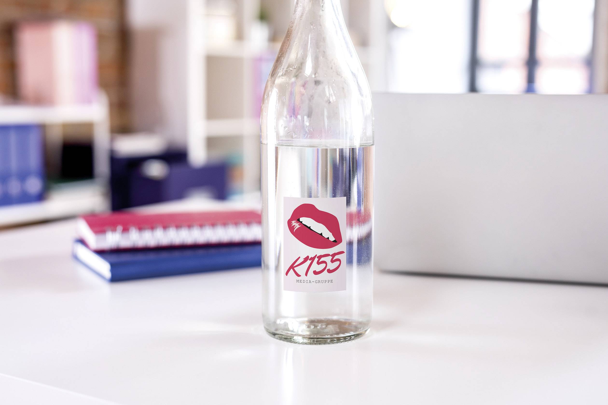 Glasflasche auf Tisch mit Etikett 'KISS' und Lippenmotiv; im Hintergrund unscharfe Bücher und Laptop; Büroambiente.