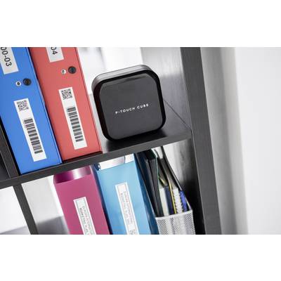 Ein schwarzes quadratisches Gerät mit der Aufschrift 'P-Touch Cube' steht in einem Regal. Daneben befinden sich farbige Ordner.