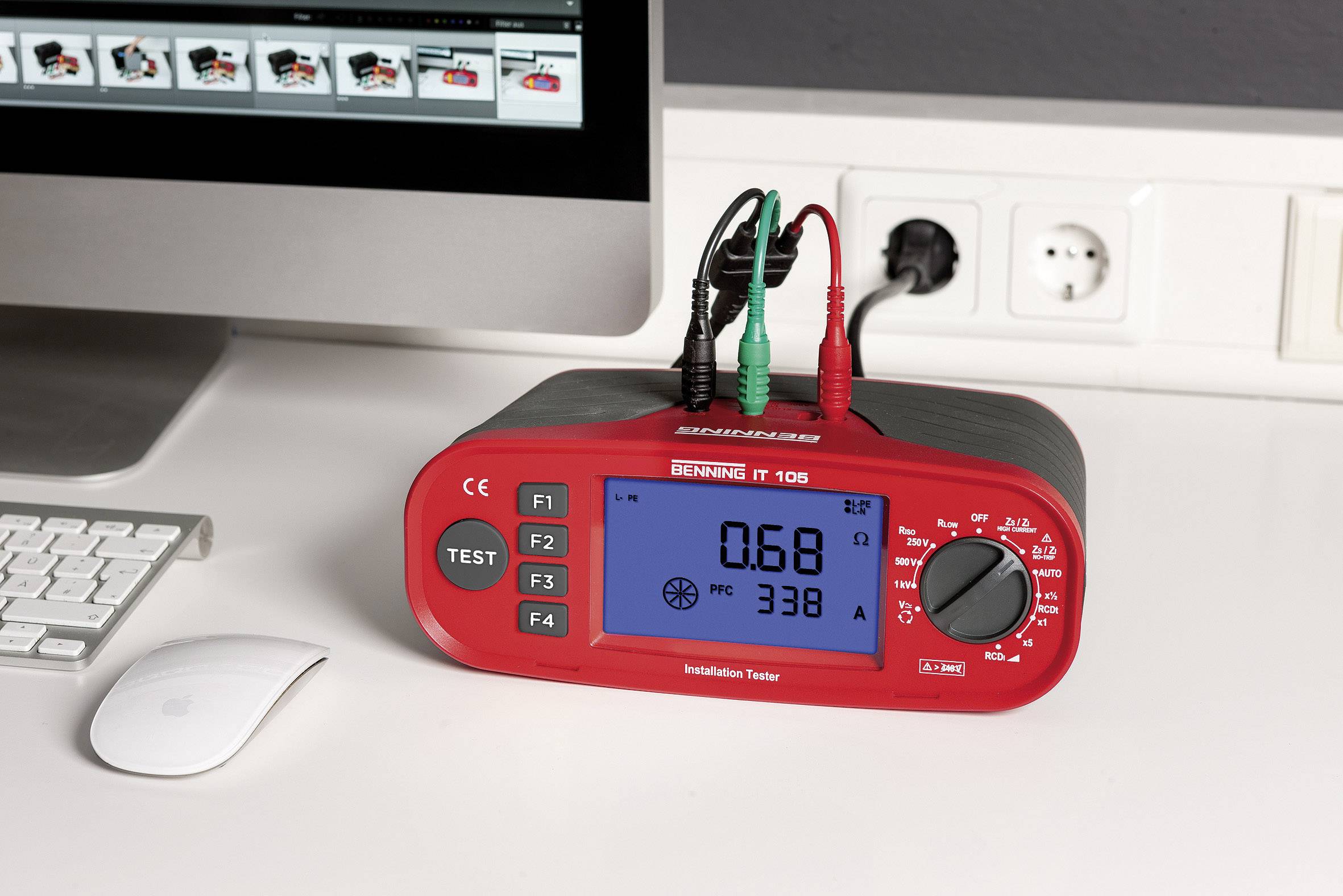 Ein rotes Installationsprüfgerät mit digitalem Display zeigt '0,68' und '338 A'. Es liegt auf einem Schreibtisch neben einem Monitor und einer Maus.